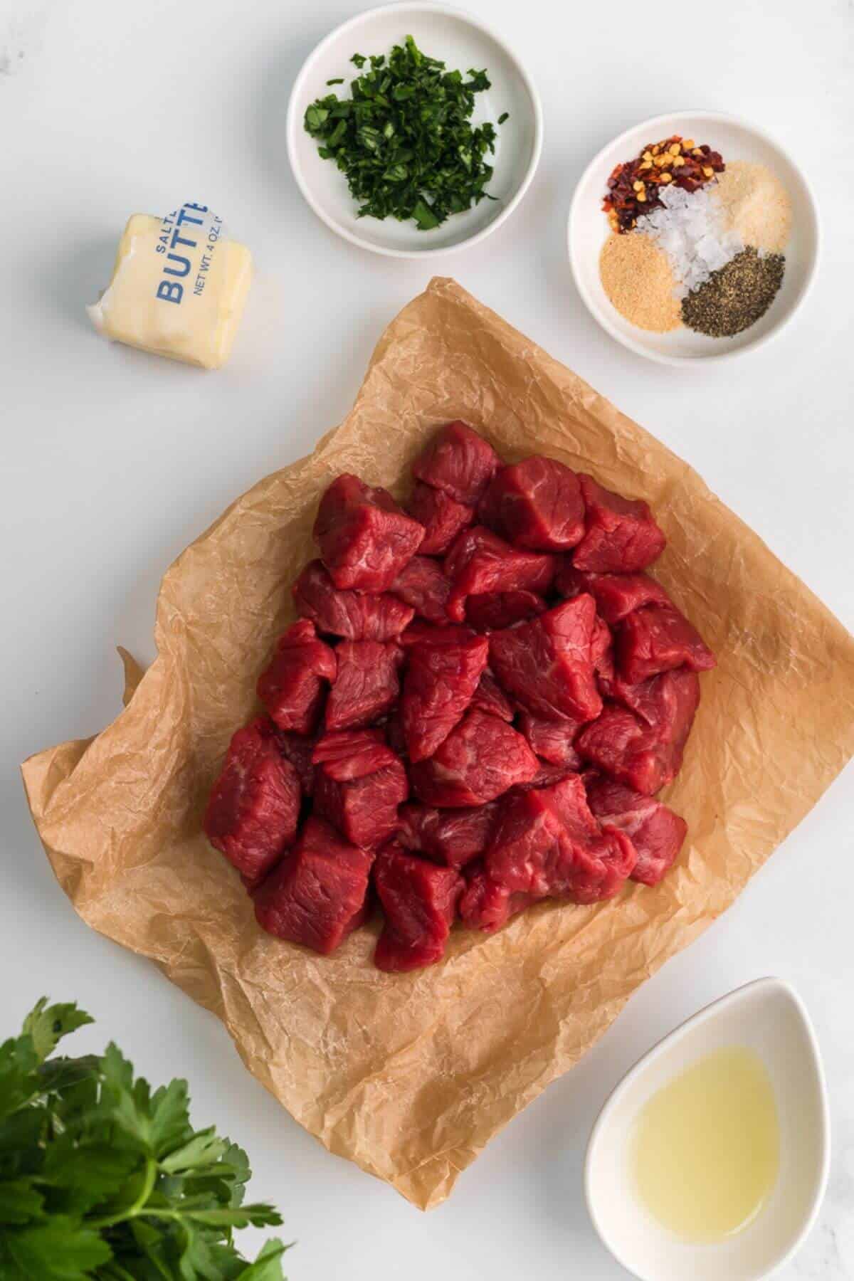 Raw beef cubes on parchment paper, surrounded by butter, chopped herbs, spices in small bowls, and a liquid in a white dish on a white background.