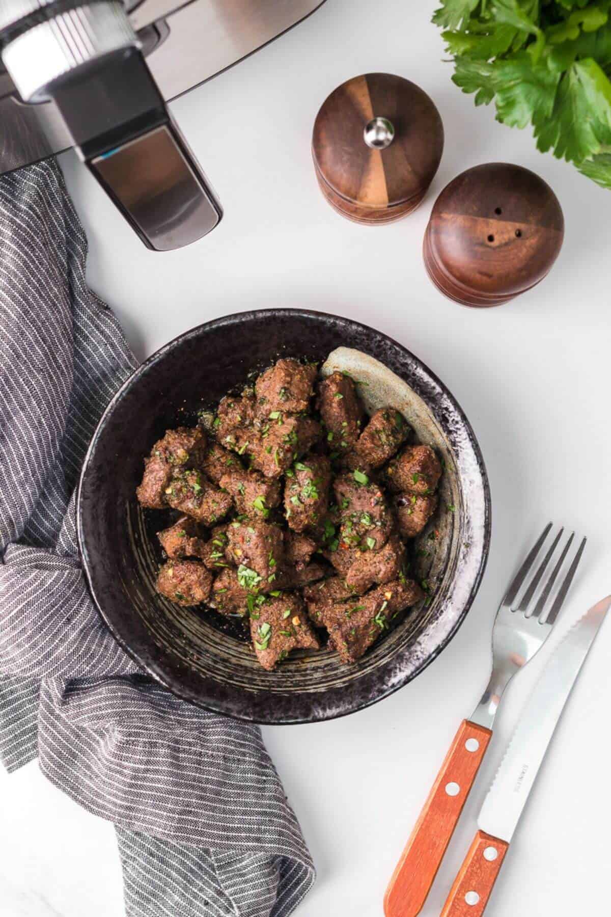 A bowl of cooked beef cubes garnished with herbs, next to a fork and knife with wooden handles, a striped napkin, salt and pepper shakers, and green leafy garnish.