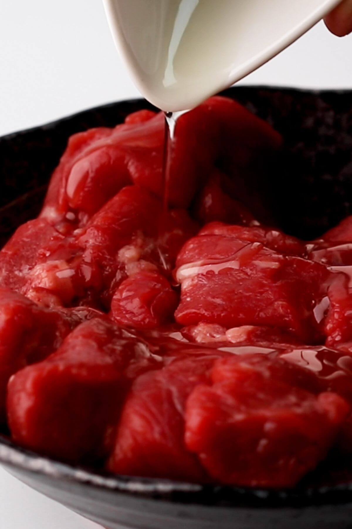 Oil pouring onto raw, cubed red meat in a dark bowl.