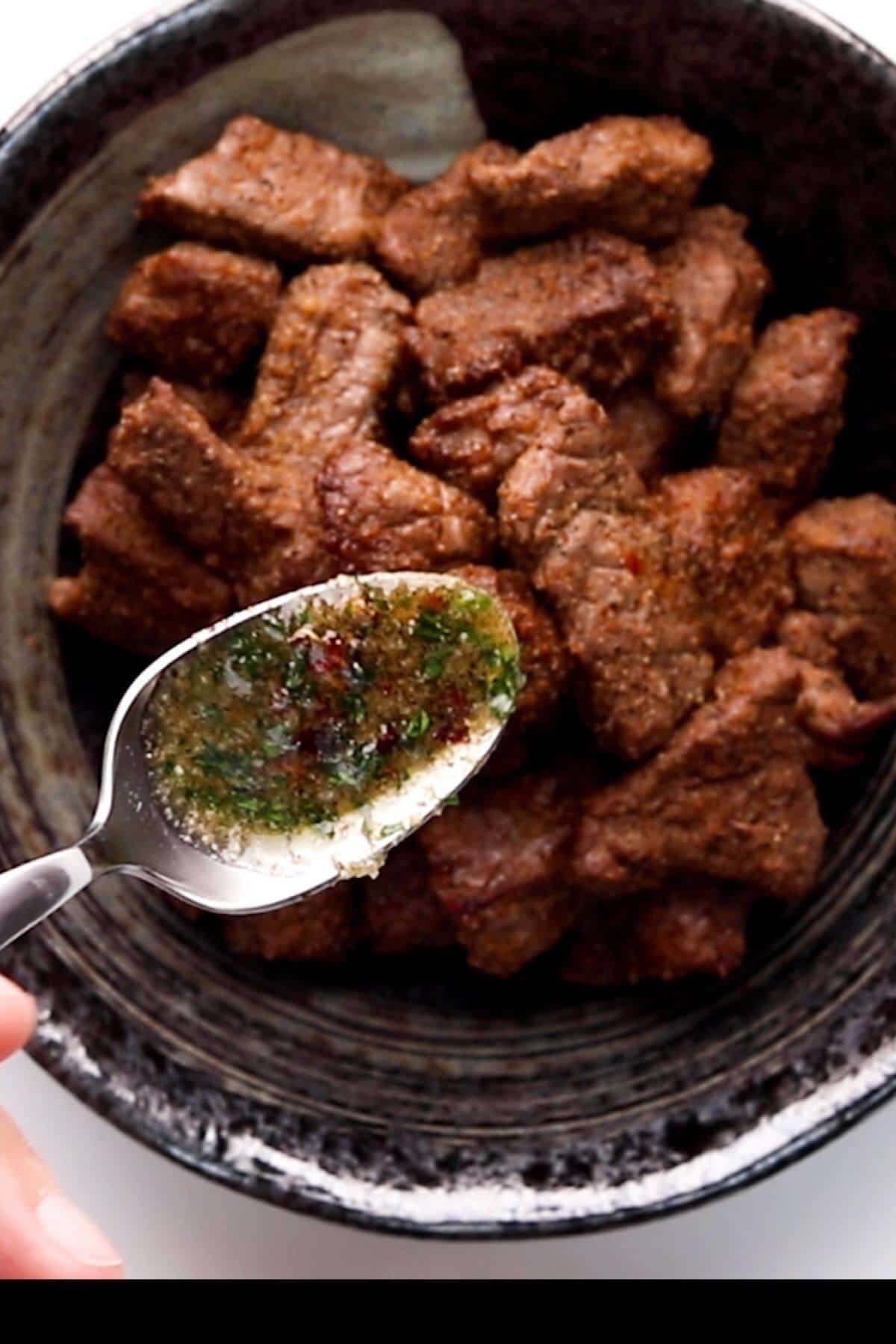 A bowl of cooked beef cubes with a spoon holding chimichurri sauce above it.