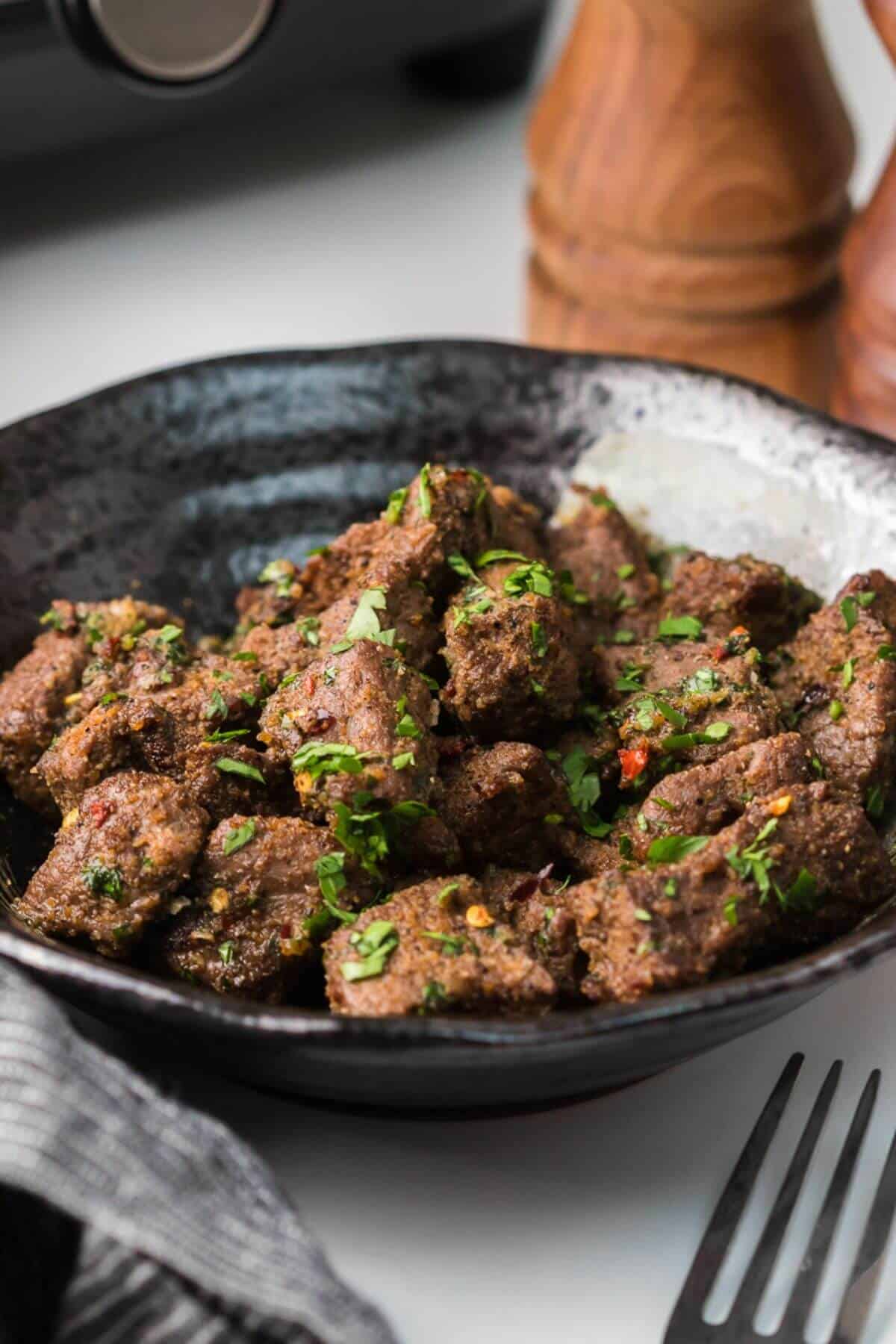 A black bowl filled with seasoned, cooked meat pieces, garnished with chopped herbs. A fork and napkin are on the side.
