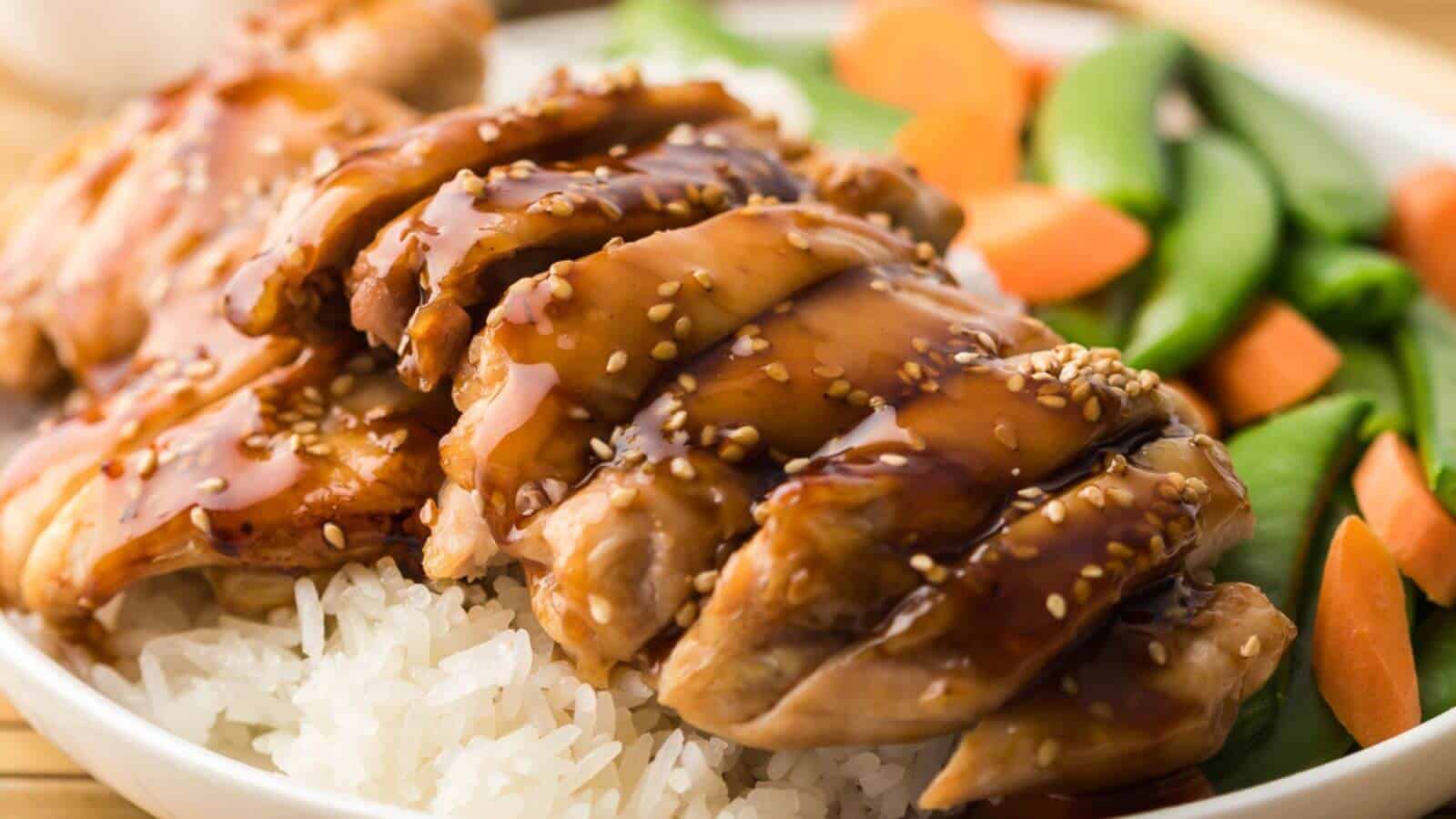 Plate of sliced teriyaki chicken over white rice, topped with sesame seeds, served with snap peas and carrot slices.