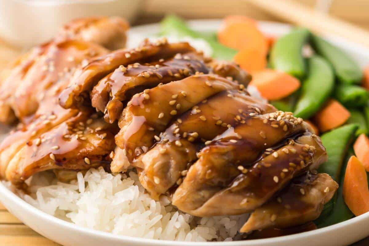 Plate of sliced teriyaki chicken topped with sesame seeds on white rice, served with snap peas and carrot slices.