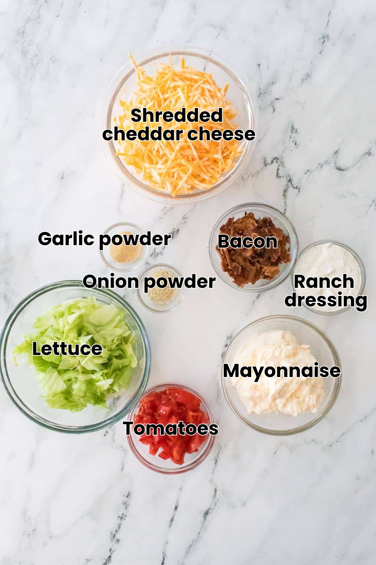 Ingredients for a salad including shredded cheese, lettuce, chopped tomatoes, bacon bits, mayonnaise, ranch dressing, onion powder, and garlic powder on a marble surface.