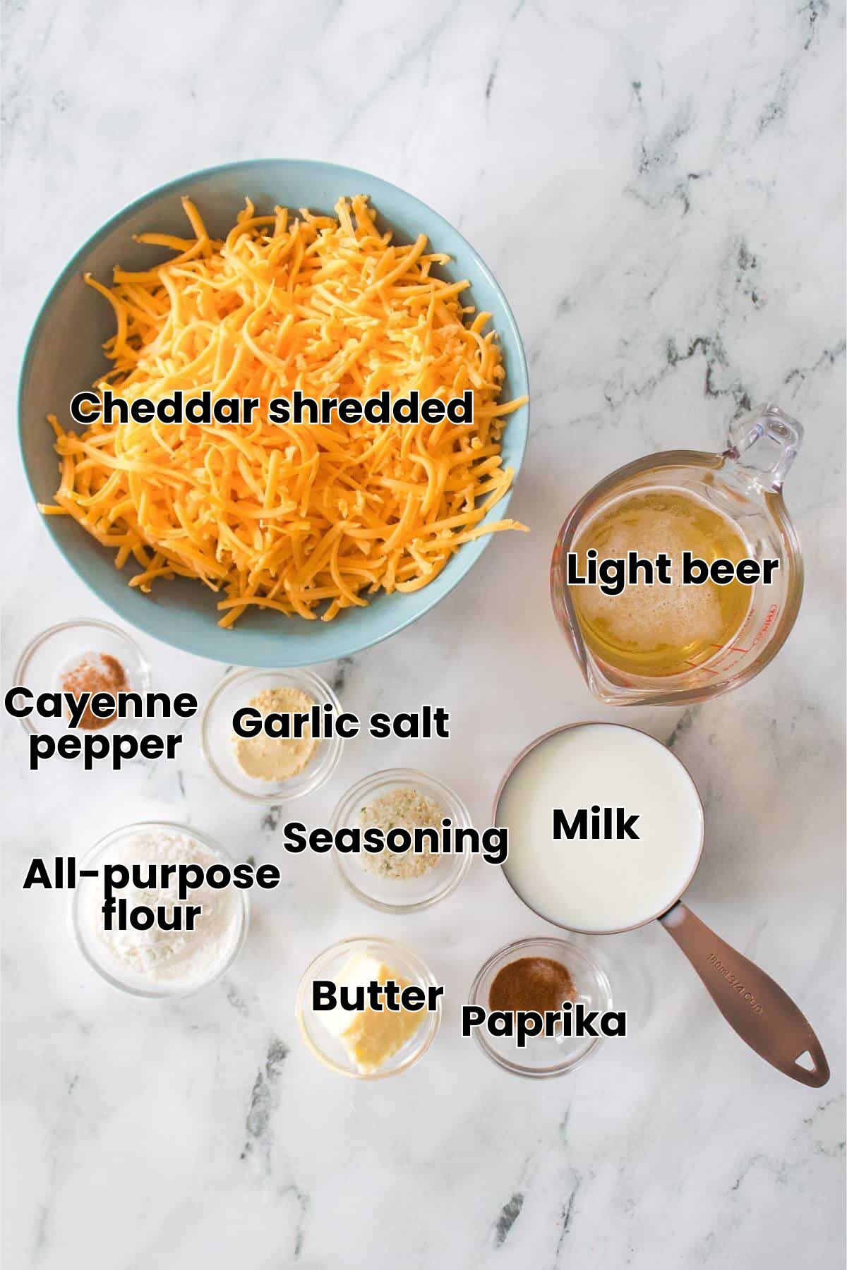A bowl of shredded cheese, a measuring cup of melted butter, and small bowls of spices, flour, and milk are arranged on a marble surface.