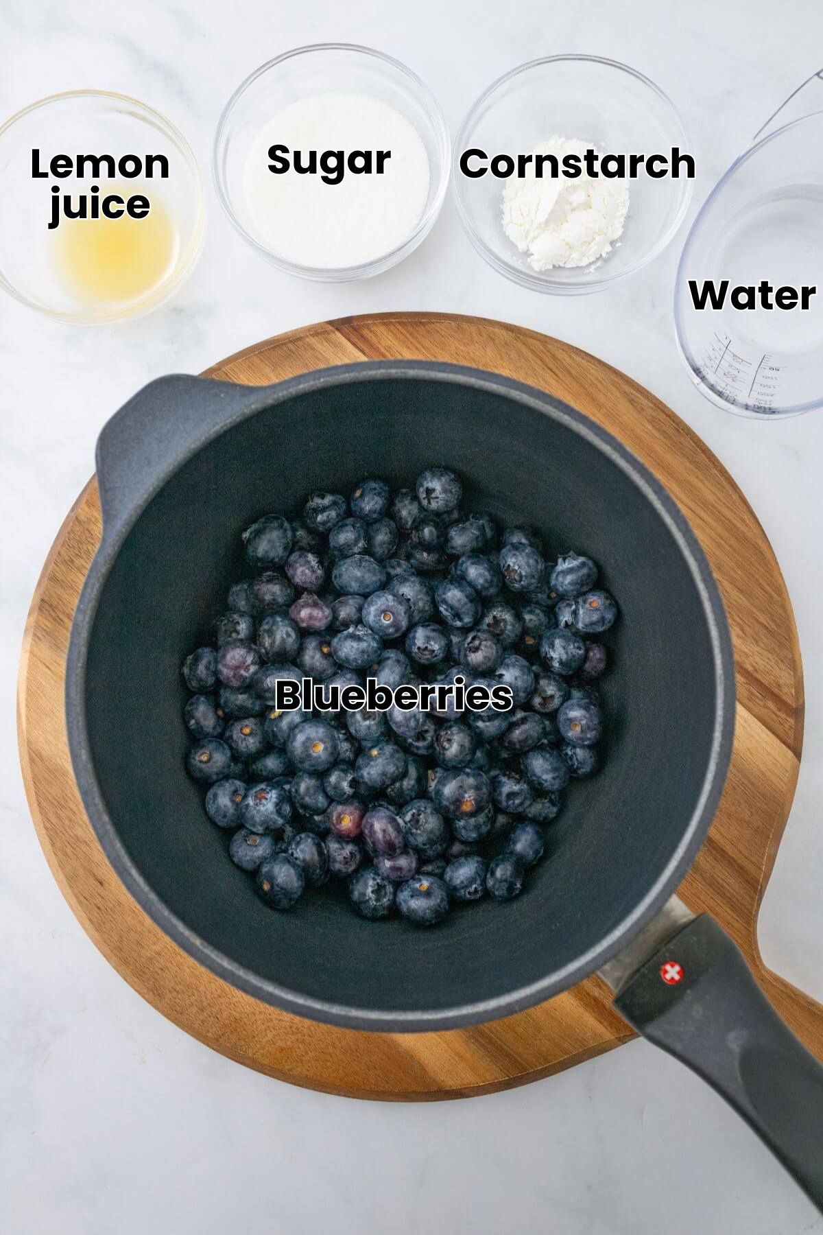 A pot filled with fresh grapes on a wooden board, surrounded by bowls of lemon juice, sugar, cornflour, and water on a marble surface.
