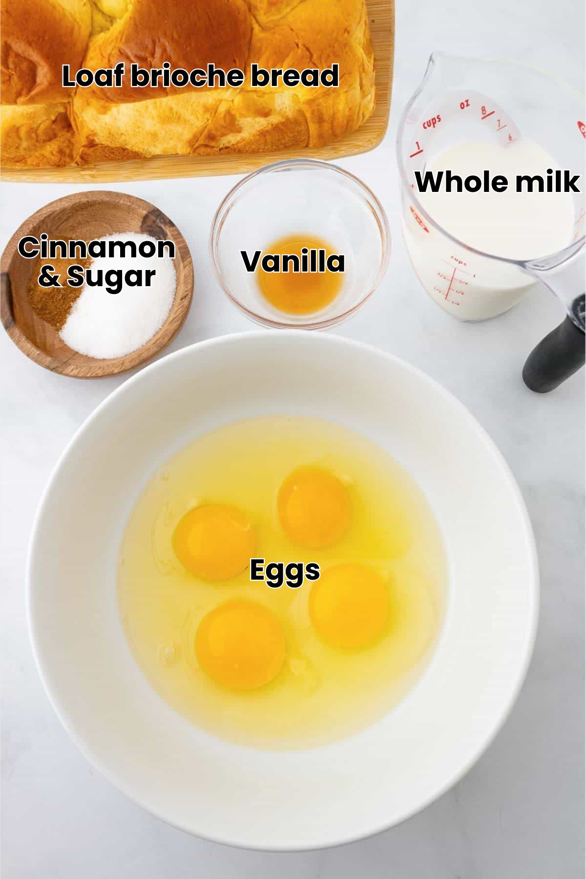 Ingredients for a recipe: three eggs in a bowl, a measuring cup of milk, small bowls of vanilla and sugar, cinnamon, and a loaf of bread.