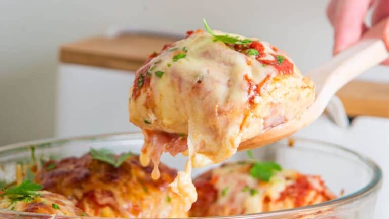 A wooden spoon holds a piece of cheesy lasagna above a glass baking dish, garnished with fresh parsley.