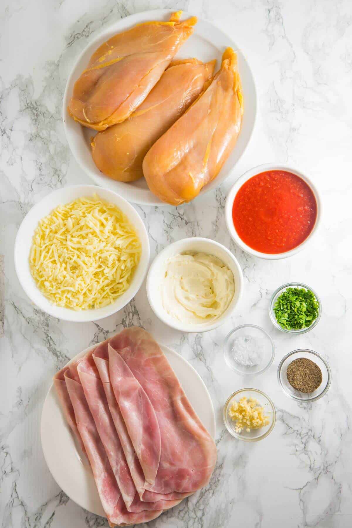 Ingredients on a marble surface: chicken breasts, tomato sauce, mozzarella cheese, ham slices, garlic, parsley, cream, salt, and pepper in separate bowls.