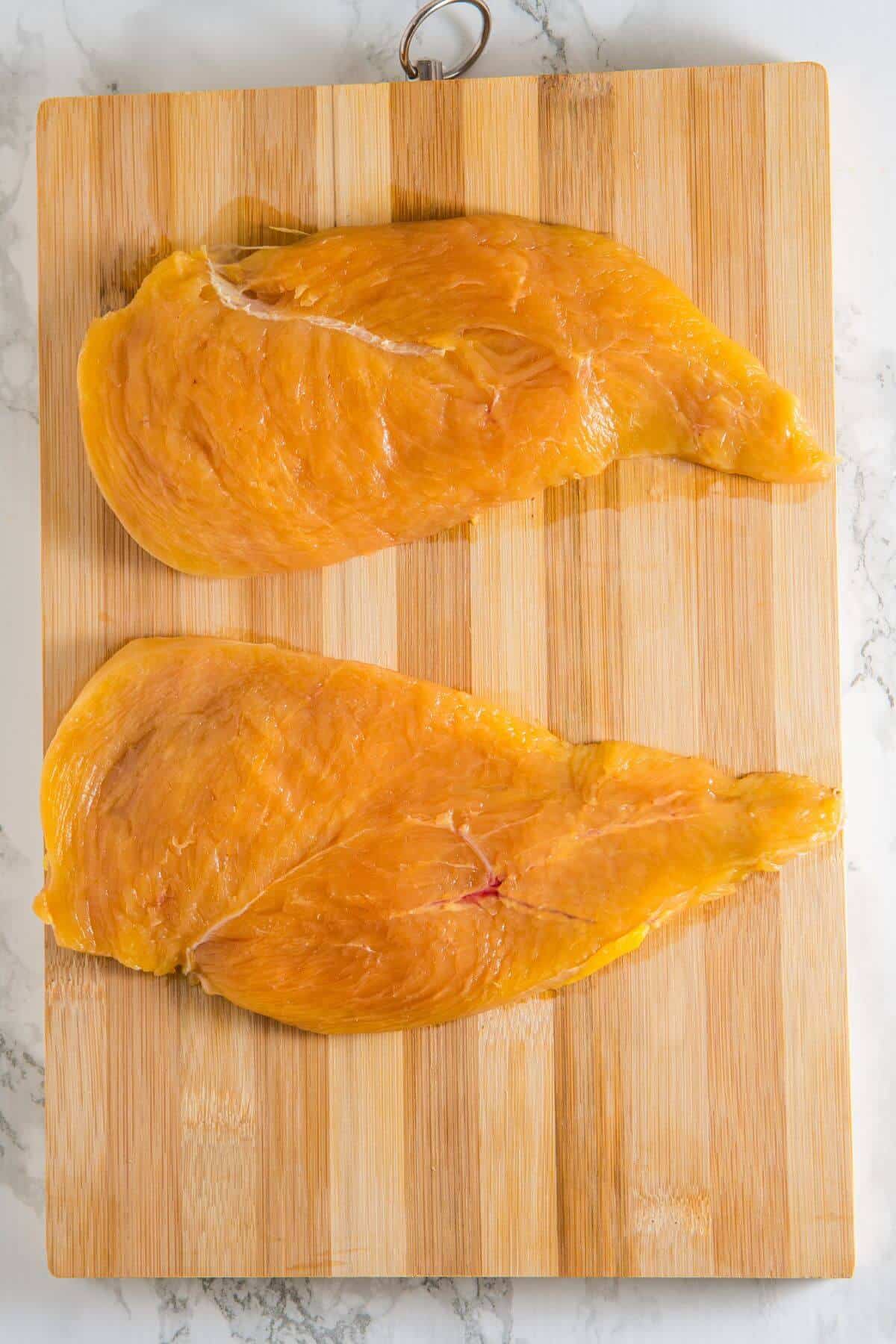 Two raw chicken breasts on a wooden cutting board placed on a marble surface.