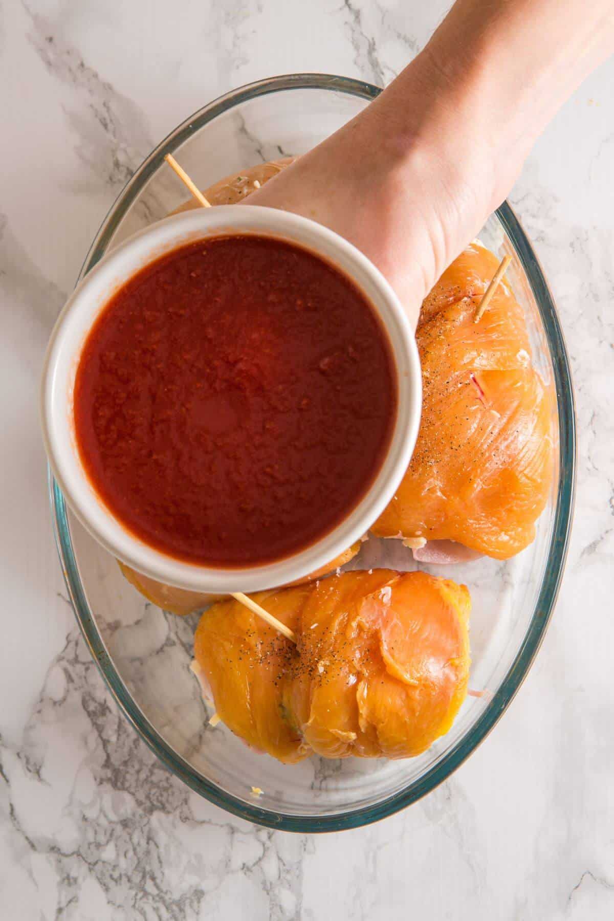 A hand holds a bowl of red sauce over a plate with three uncooked, seasoned chicken breasts secured with toothpicks, on a marble surface.