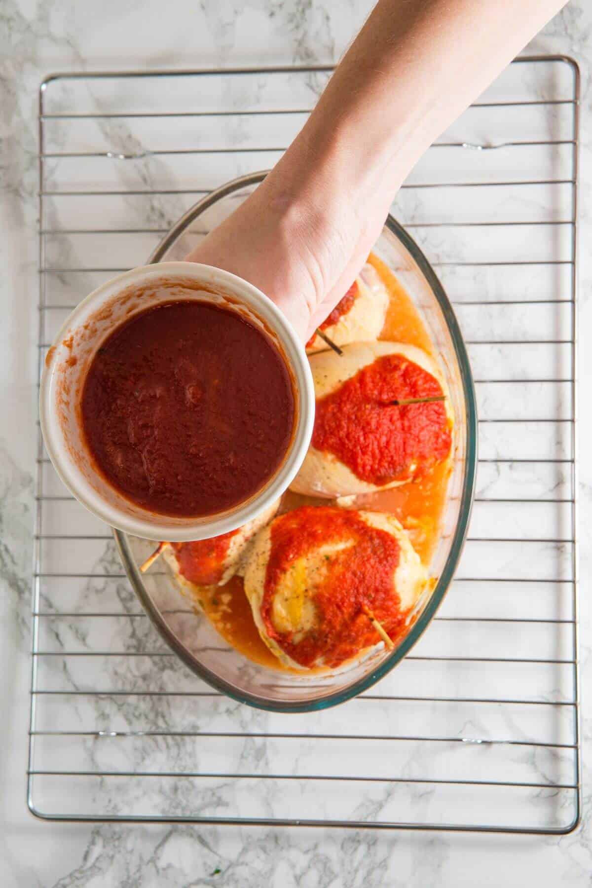 A hand pouring tomato sauce over stuffed peppers in a glass baking dish on a cooling rack.