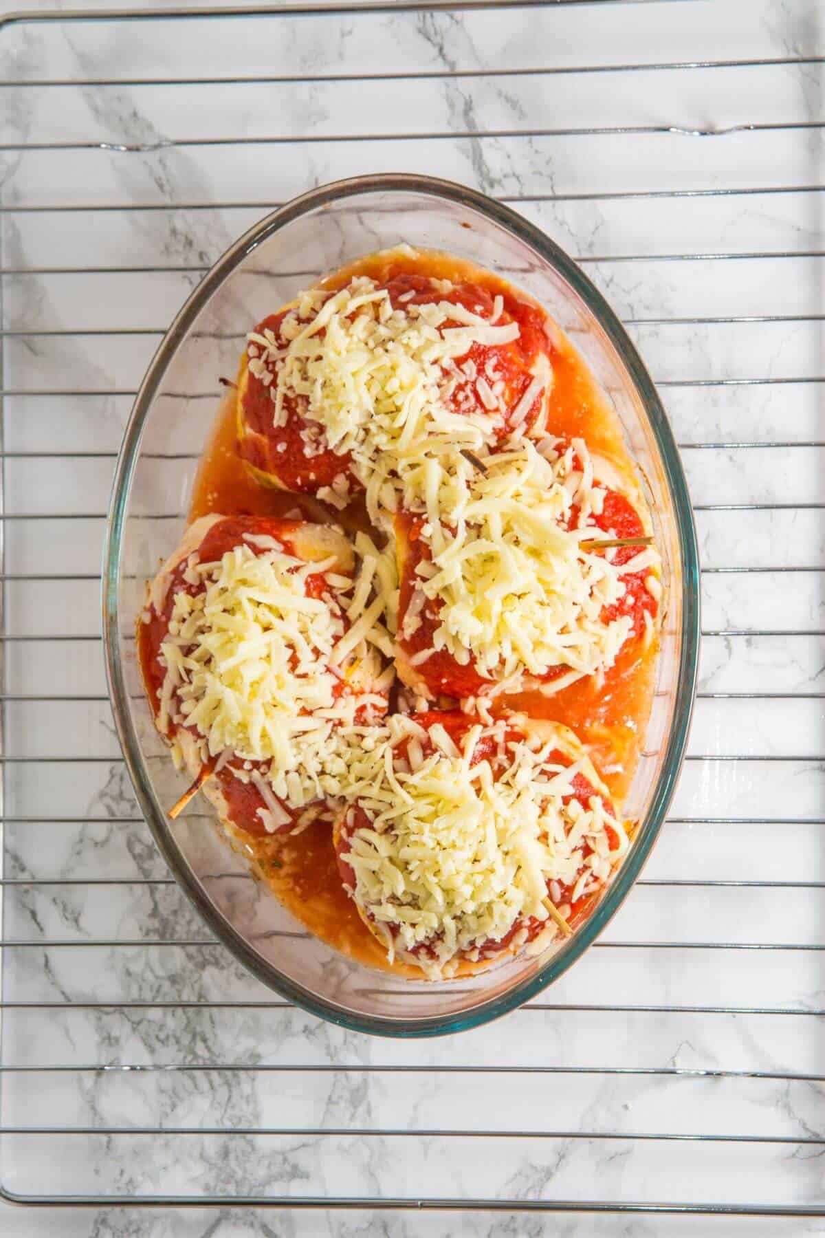 Four stuffed tomatoes topped with grated cheese in a glass baking dish on a marble and wire rack background.