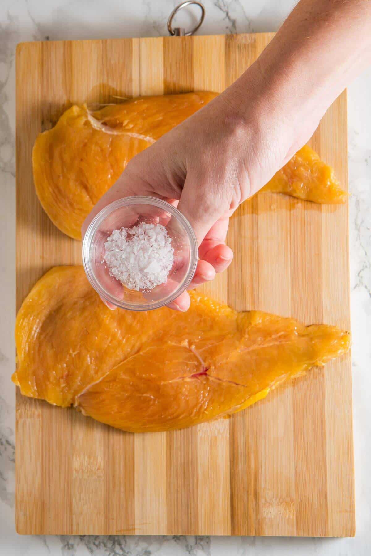 Two raw chicken breasts on a wooden cutting board with a hand holding a small bowl of salt above them.