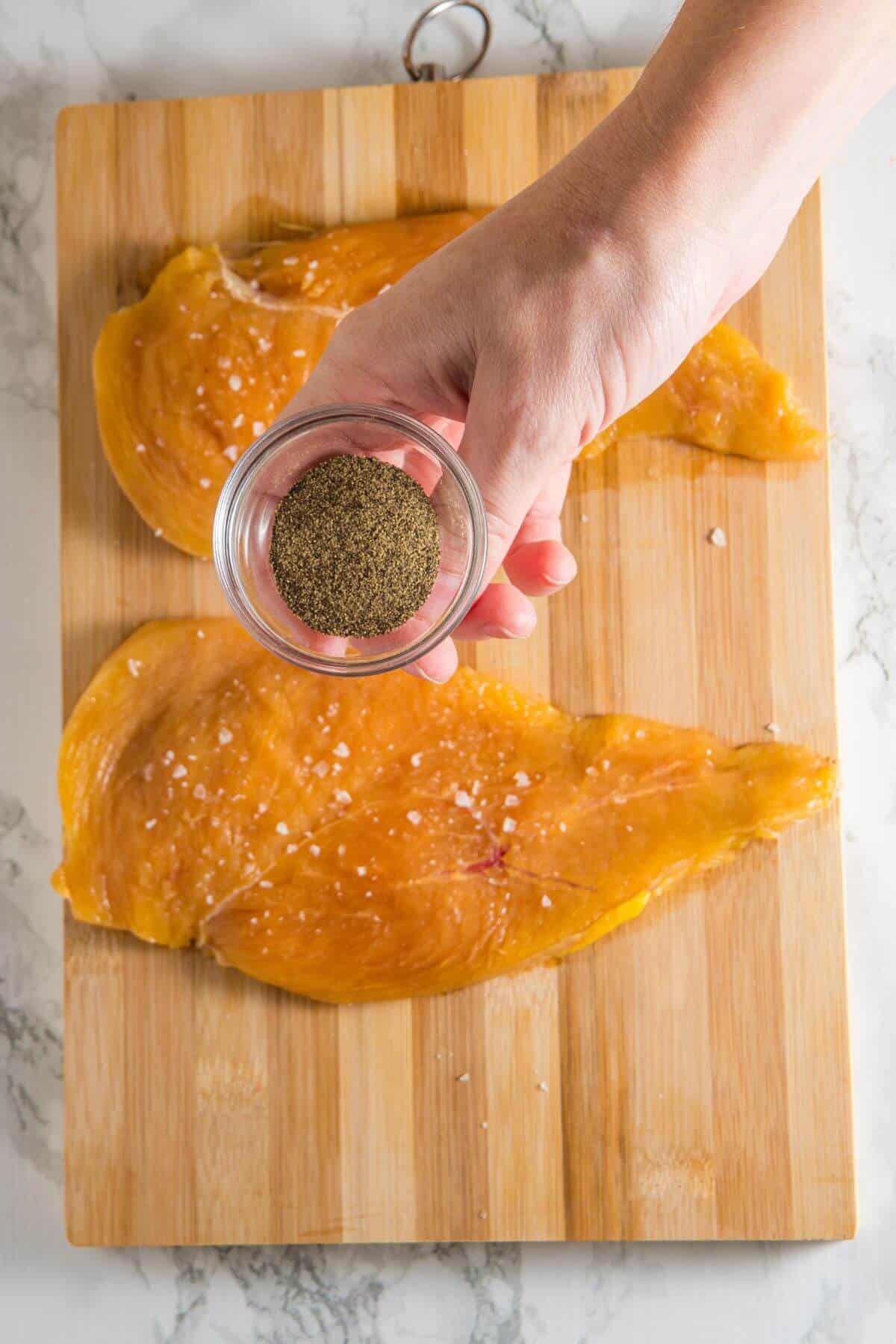 A hand holds a small glass bowl of ground pepper above two seasoned chicken breasts on a wooden cutting board.