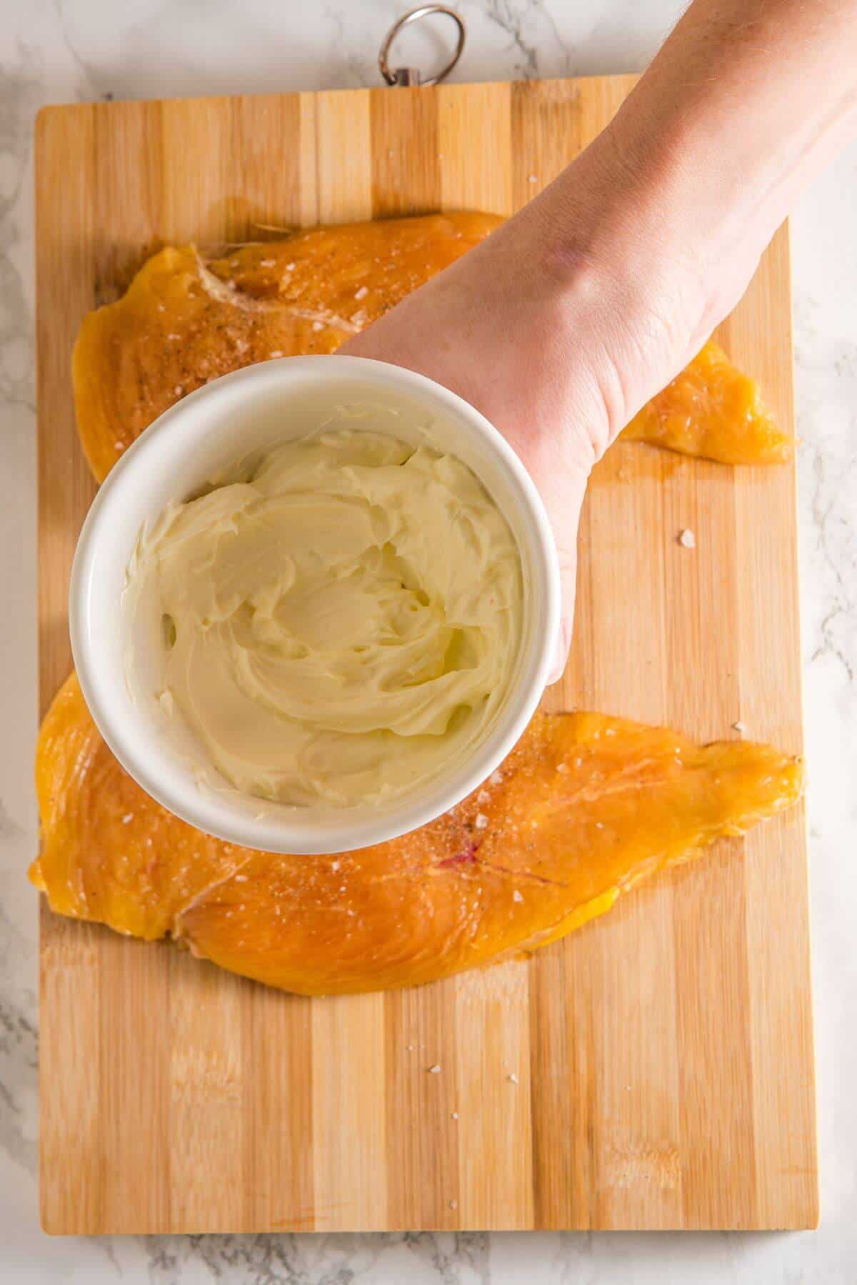 A hand holds a white bowl of cream cheese above two seasoned chicken breasts on a wooden cutting board.