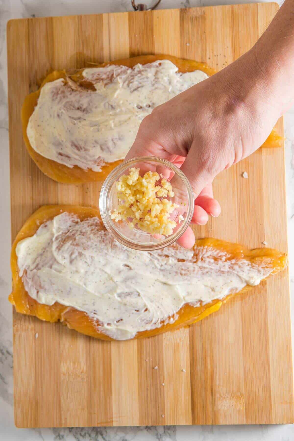 A hand holding a small bowl of minced garlic above two pieces of chicken coated with a white sauce on a wooden board.