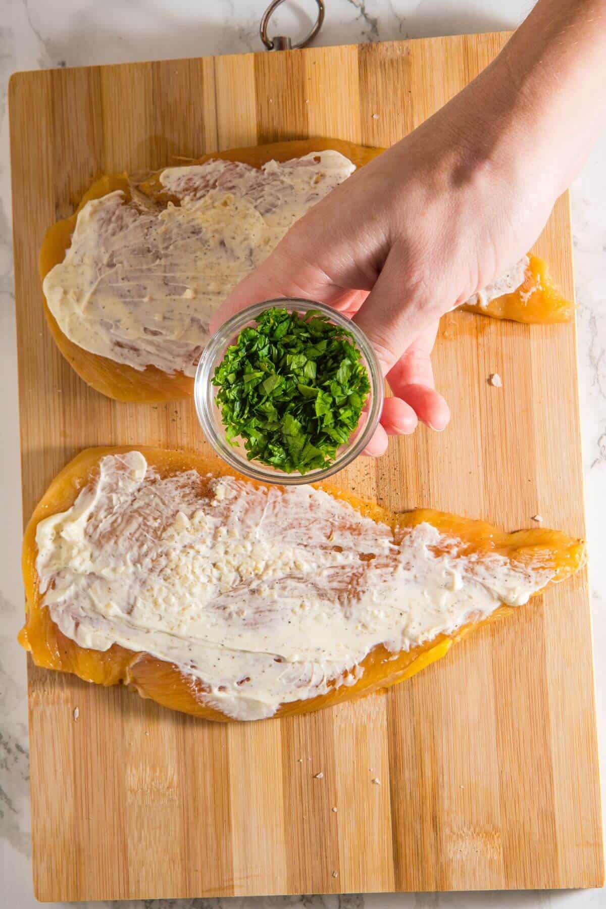 Two chicken breasts on a wooden board are spread with cream sauce. A hand holds a small bowl of chopped green herbs above them.