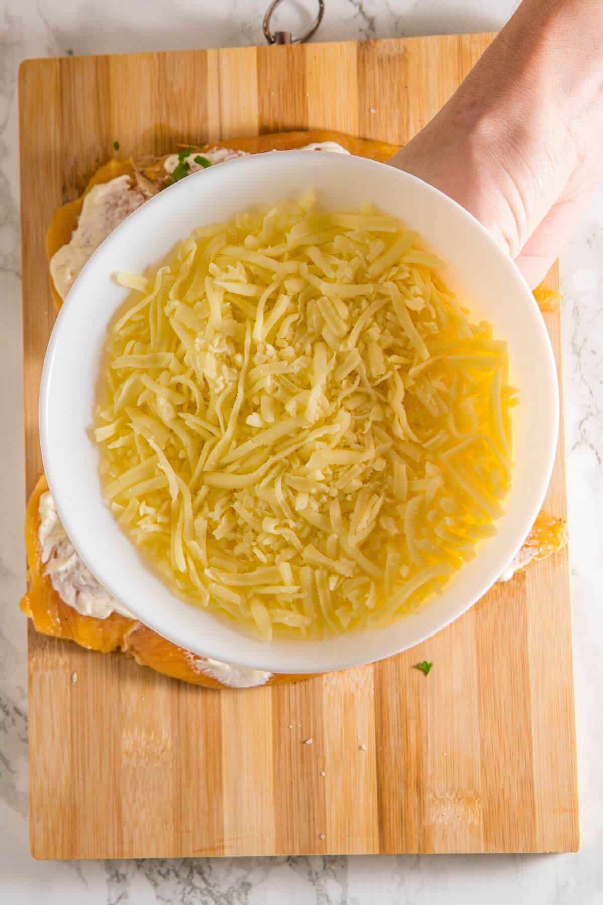 A hand holds a white bowl filled with grated cheese over a wooden cutting board.