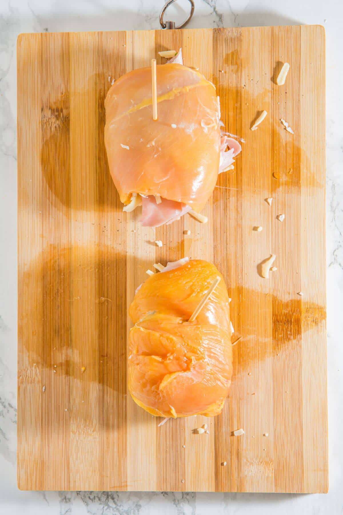 Two uncooked chicken breasts stuffed with ham and cheese, secured with toothpicks, placed on a wooden cutting board.