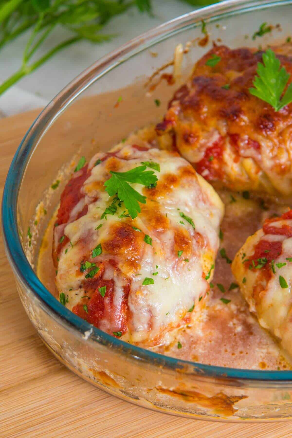 A glass baking dish with baked cheesy stuffed chicken breasts garnished with tomato sauce and parsley.