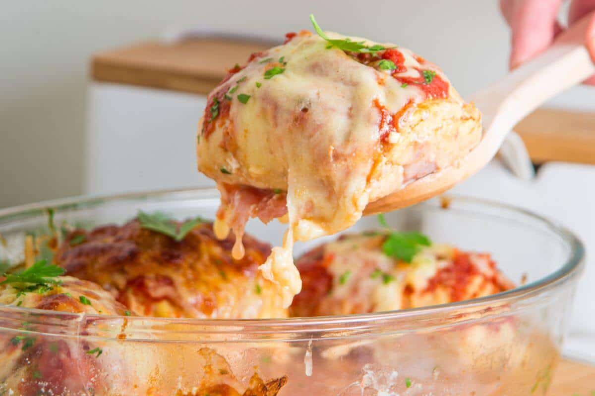 A wooden spoon lifts a portion of cheesy baked chicken from a glass baking dish.