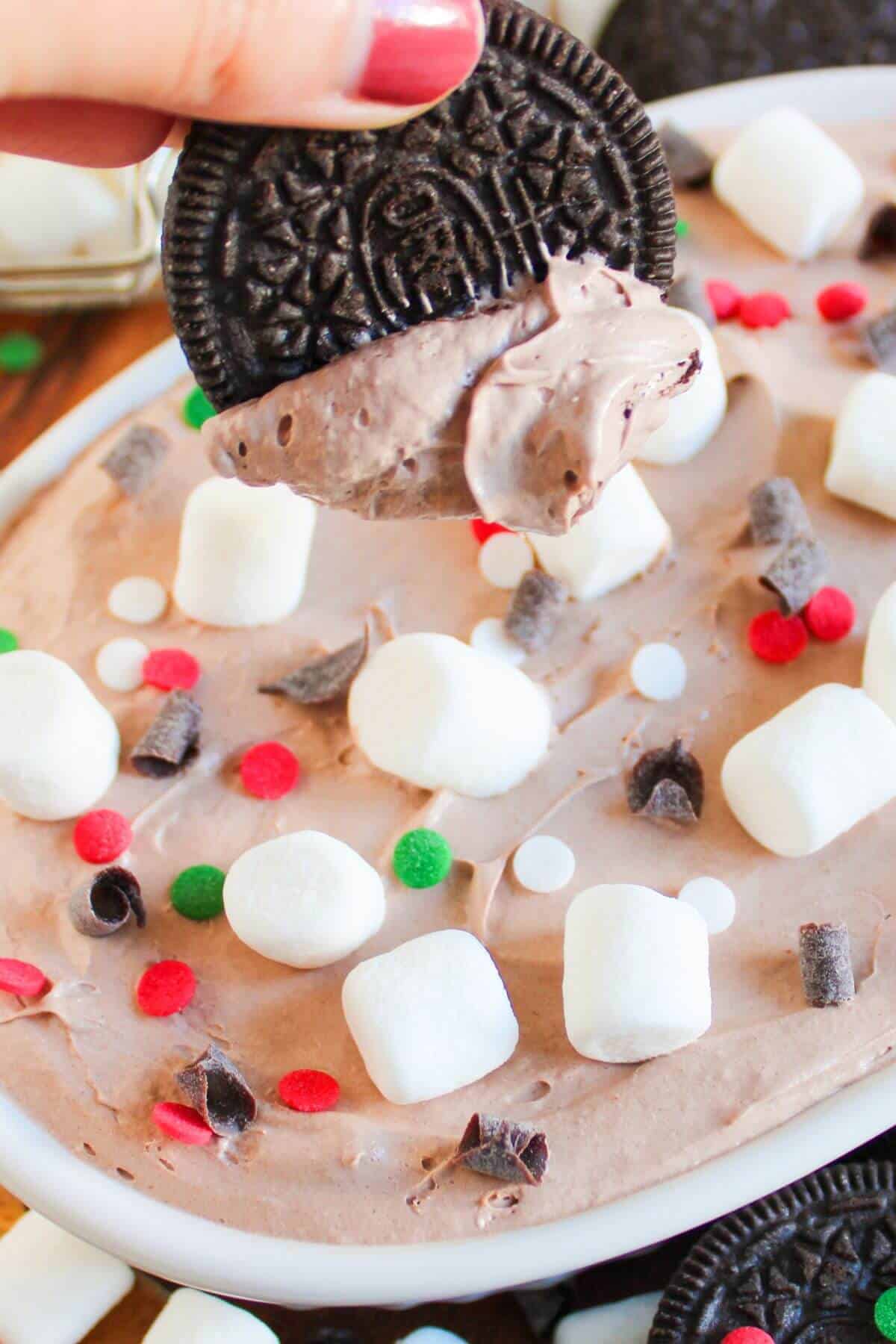 A hand holding a cream-covered Oreo above a dish of chocolate dip, topped with marshmallows, chocolate chunks, and red and green sprinkles.