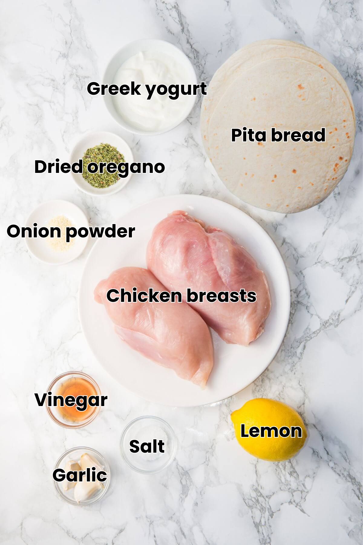 Raw chicken breasts on a plate surrounded by tortillas, lemon, yogurt, garlic, seasoning, vinegar, and oil on a marble countertop.