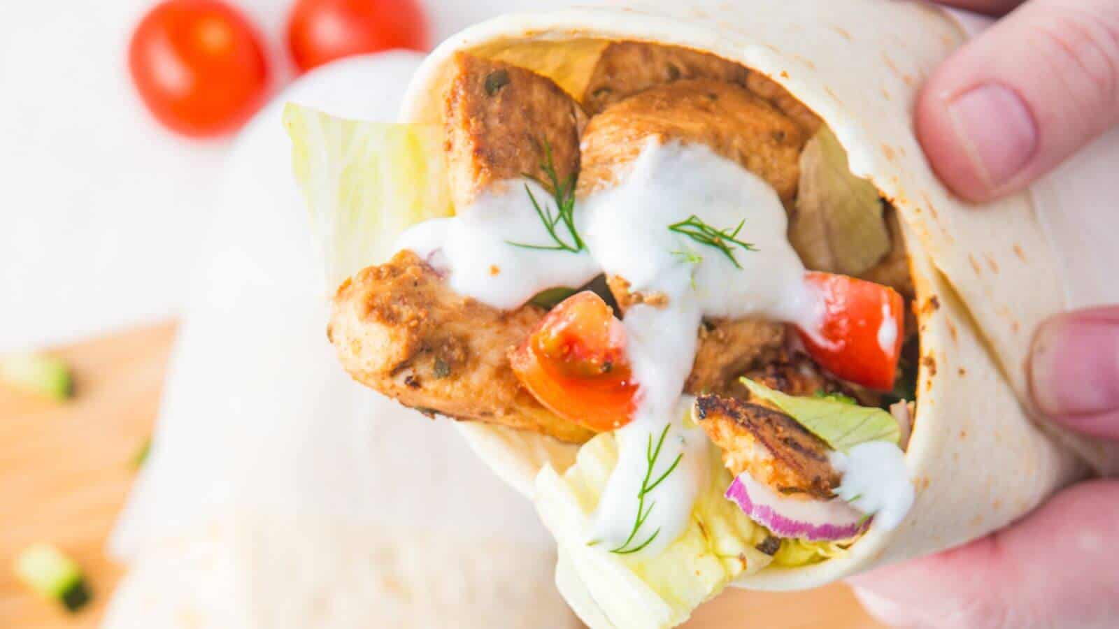 A hand holding a chicken wrap with lettuce, tomatoes, red onions, and white sauce in a tortilla.