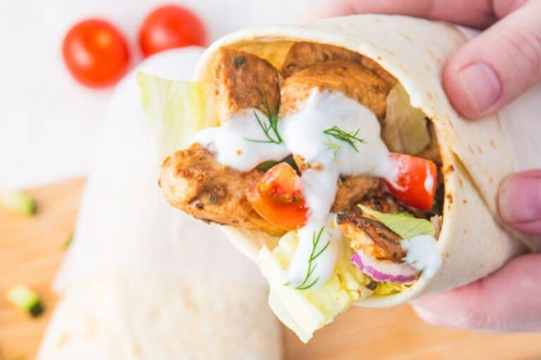 Person holding a chicken wrap with lettuce, tomatoes, and sauce, against a light background with cherry tomatoes in the back.