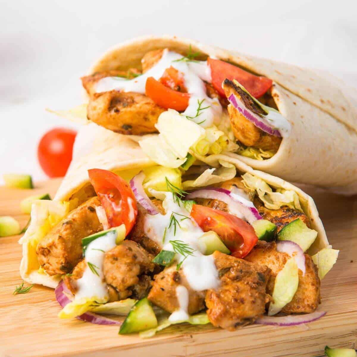Two chicken wraps filled with grilled chicken, lettuce, tomatoes, cucumbers, red onions, and drizzled with white sauce. Placed on a wooden cutting board.