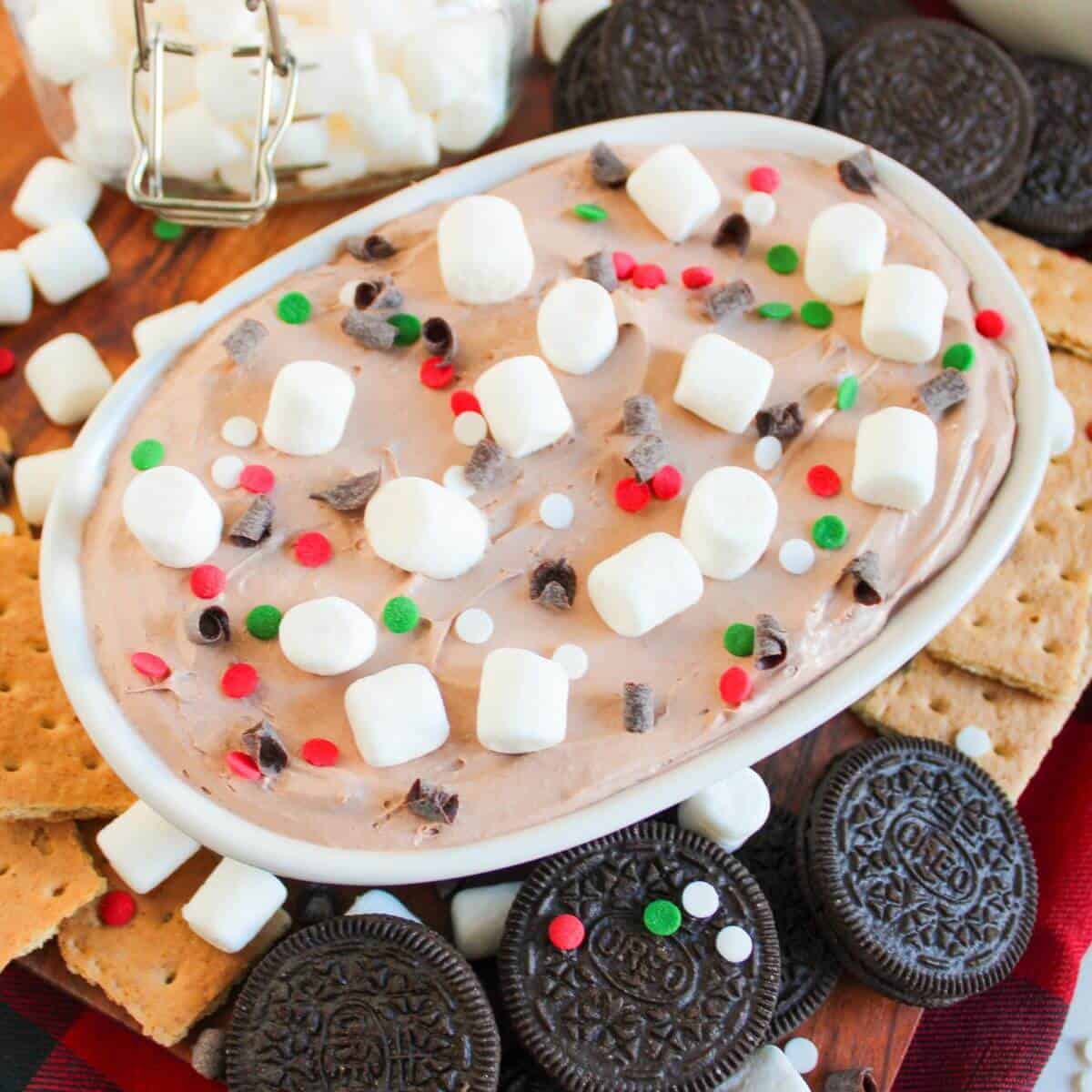 A festive dessert dip topped with mini marshmallows, chocolate chips, and sprinkles in a dish. Surrounded by cookies and graham crackers.
