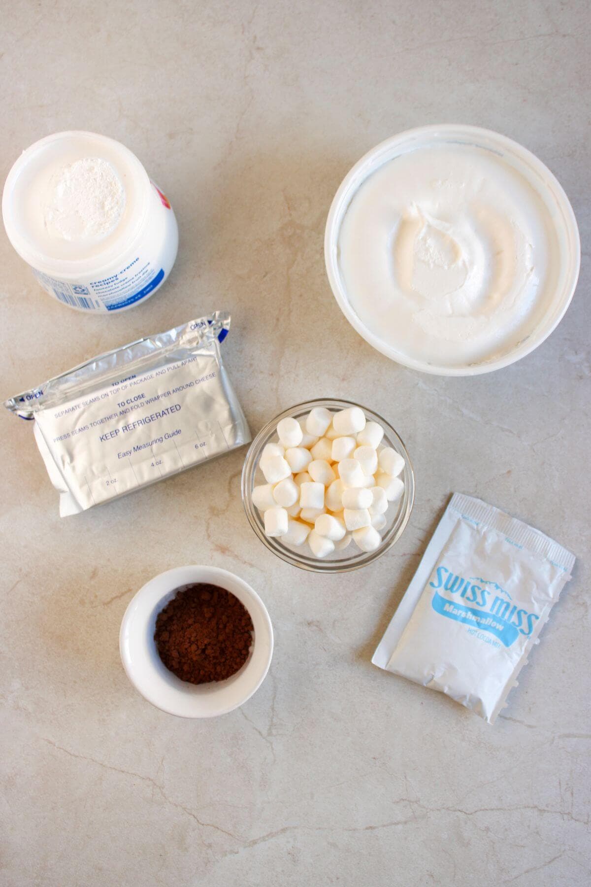 Ingredients for a dessert are arranged on a countertop, including whipped topping, mini marshmallows, hot cocoa mix, a white powder, pudding mix, and cocoa powder.