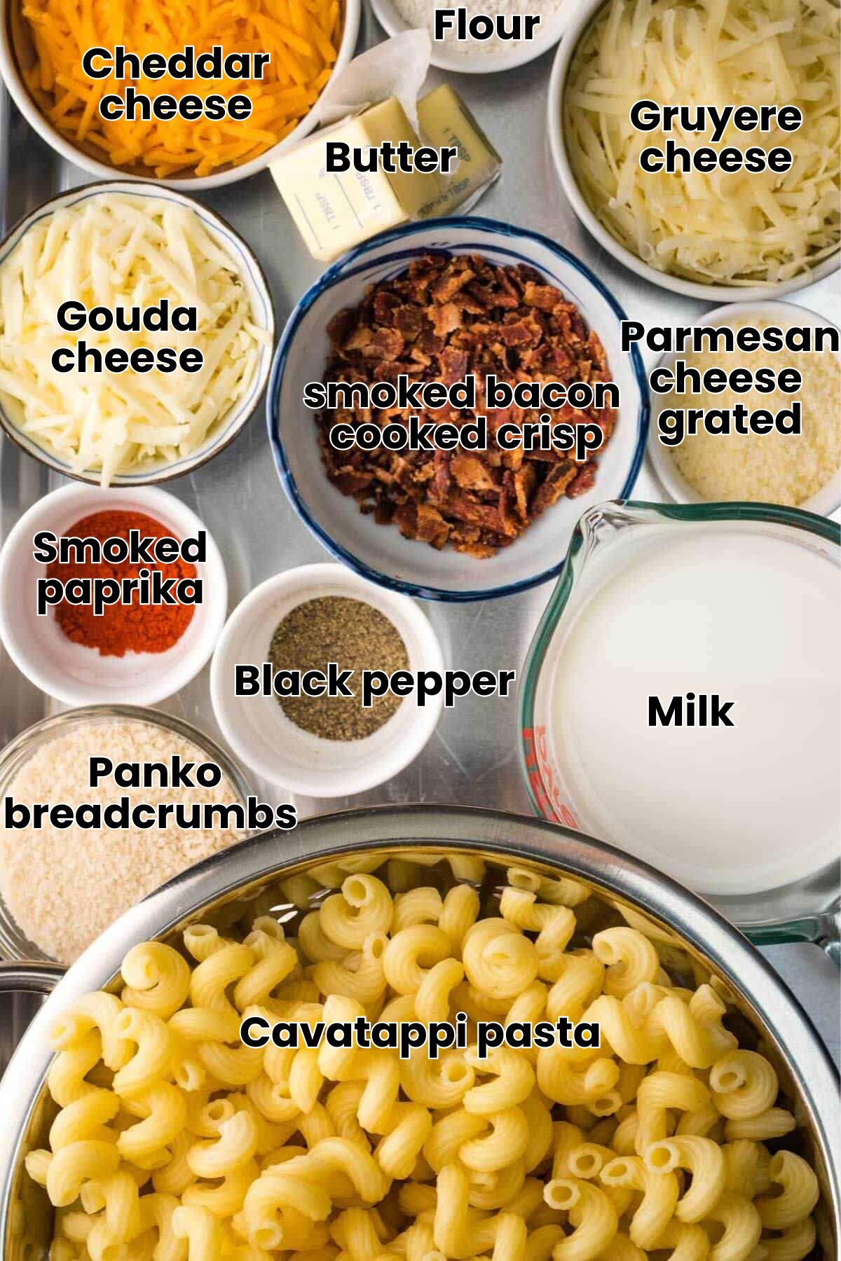 Ingredients for macaroni and cheese are arranged on a table, including pasta, milk, bacon, shredded cheese, butter, and various spices in bowls.