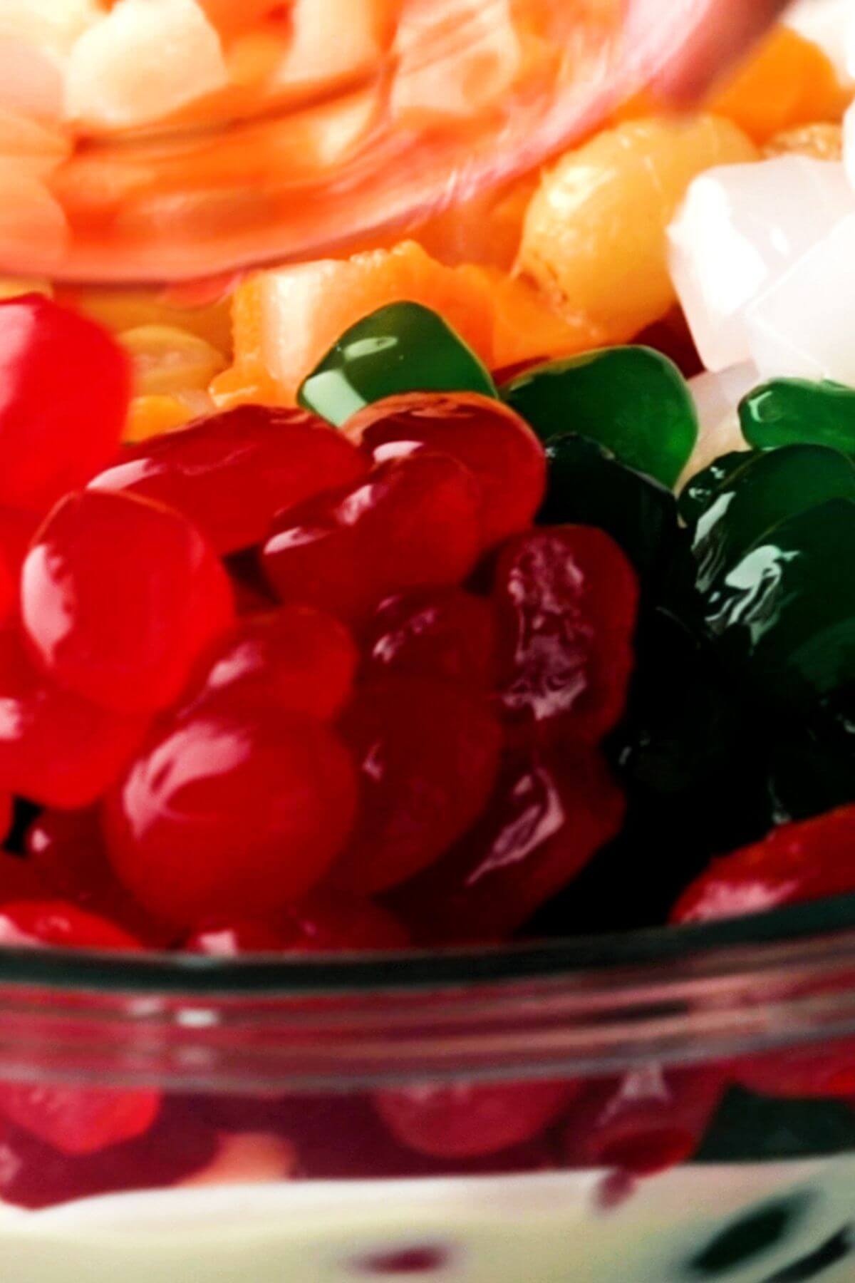 A close-up of a dessert bowl with colorful jelly cubes in red, green, and orange, alongside white cubes, possibly coconut or lychee, mixed together.