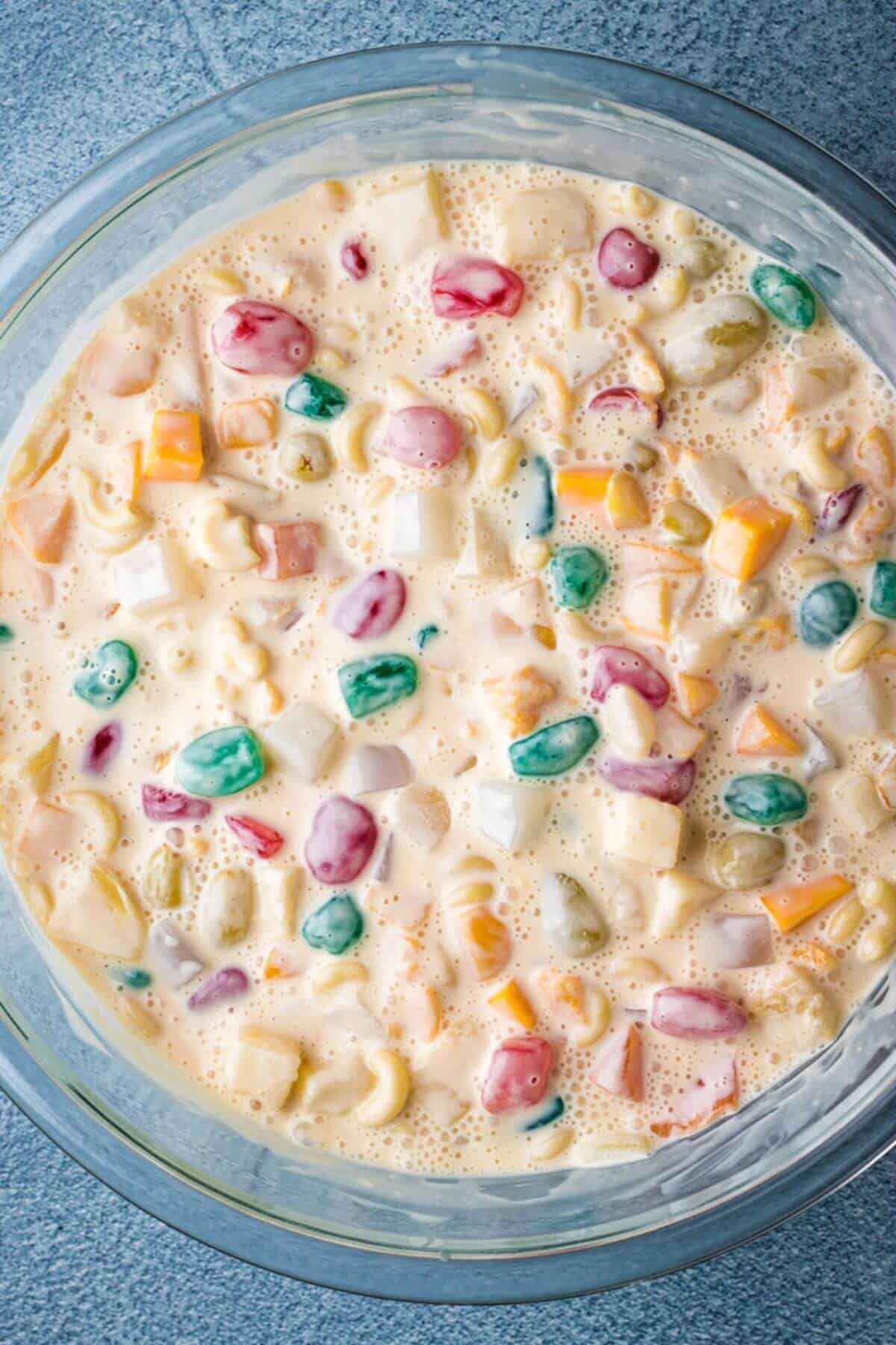 A bowl of creamy fruit salad with colorful marshmallows, small pasta, and diced fruits in a rich sauce.