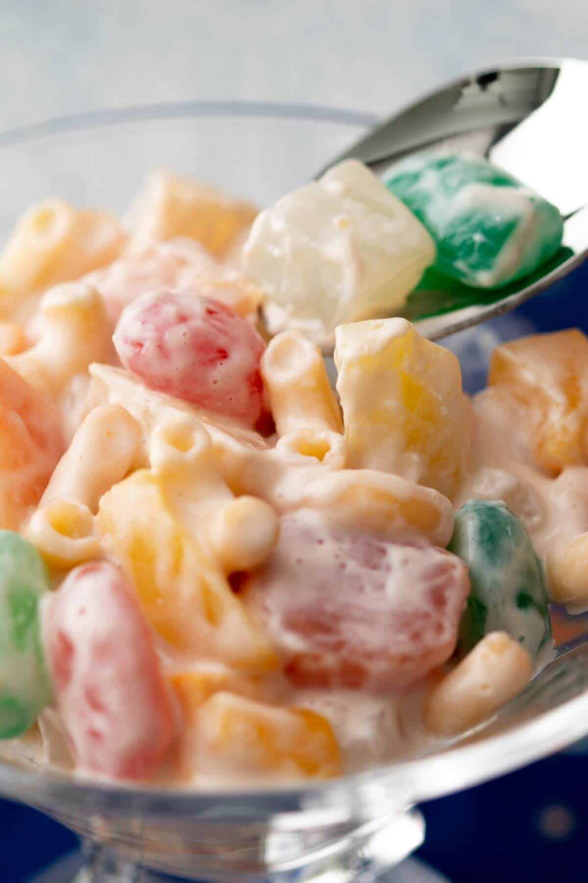 A glass bowl of fruit salad with colorful diced fruit and creamy dressing, and a spoon scooping it.