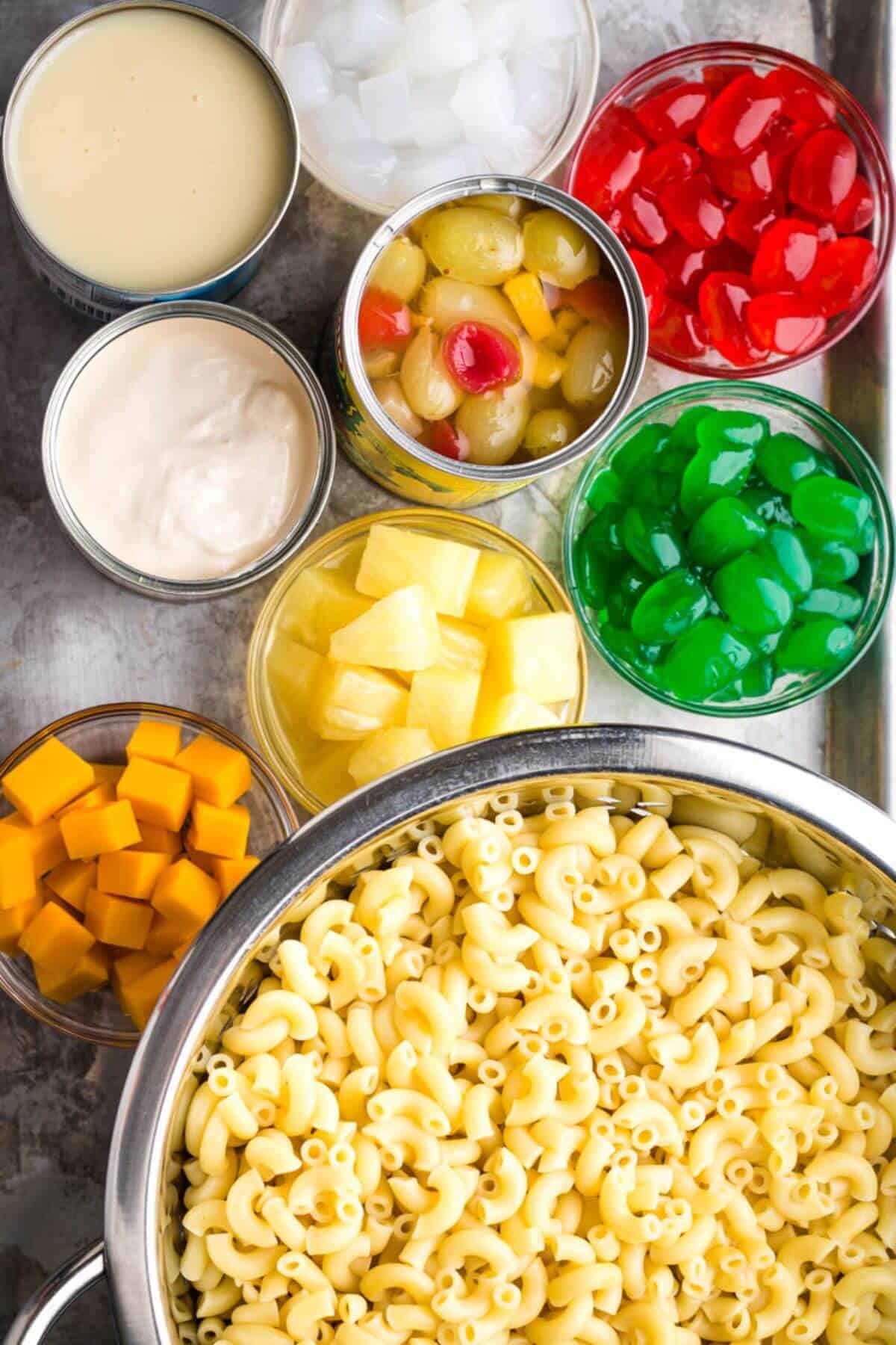 Ingredients for a dessert salad: macaroni in a pot, bowls of cheese cubes, sweetened condensed milk, fruit cocktail, diced pineapple, maraschino cherries, green cherries, and diced coconut.