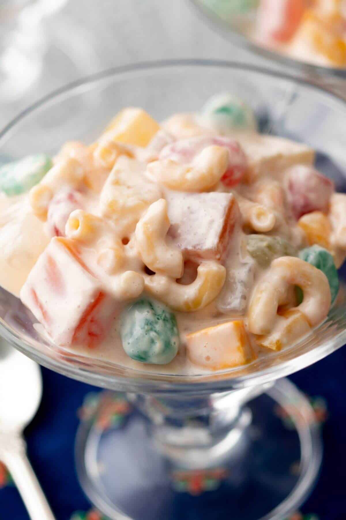 A glass bowl filled with macaroni salad mixed with colorful fruits and creamy dressing. A spoon is partially visible in the background.