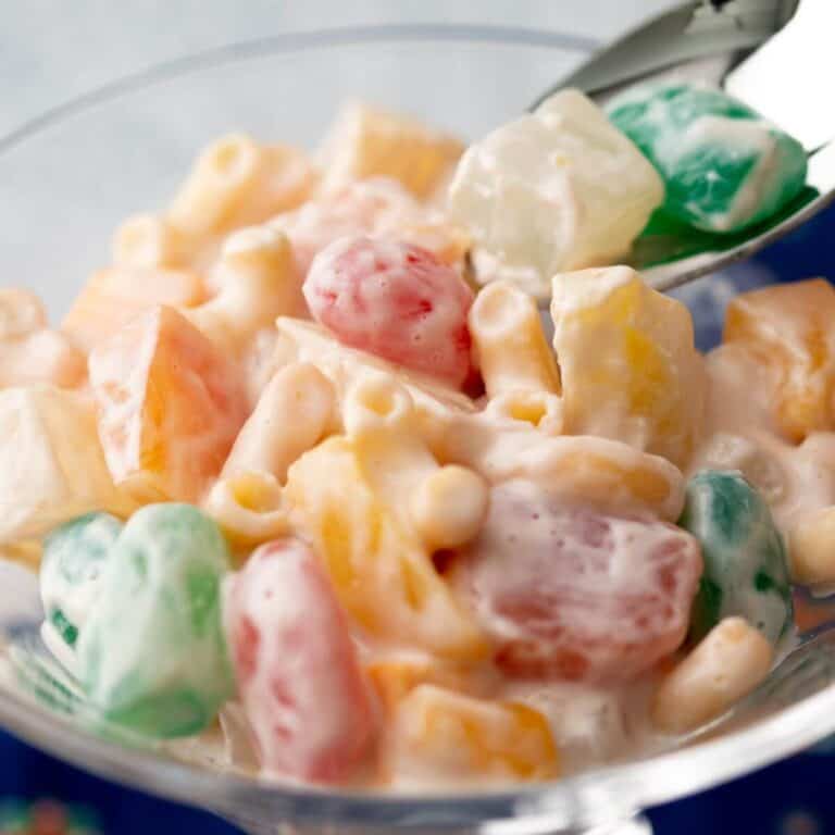 A glass bowl filled with a colorful fruit salad mixed with creamy dressing and a spoon beside it.