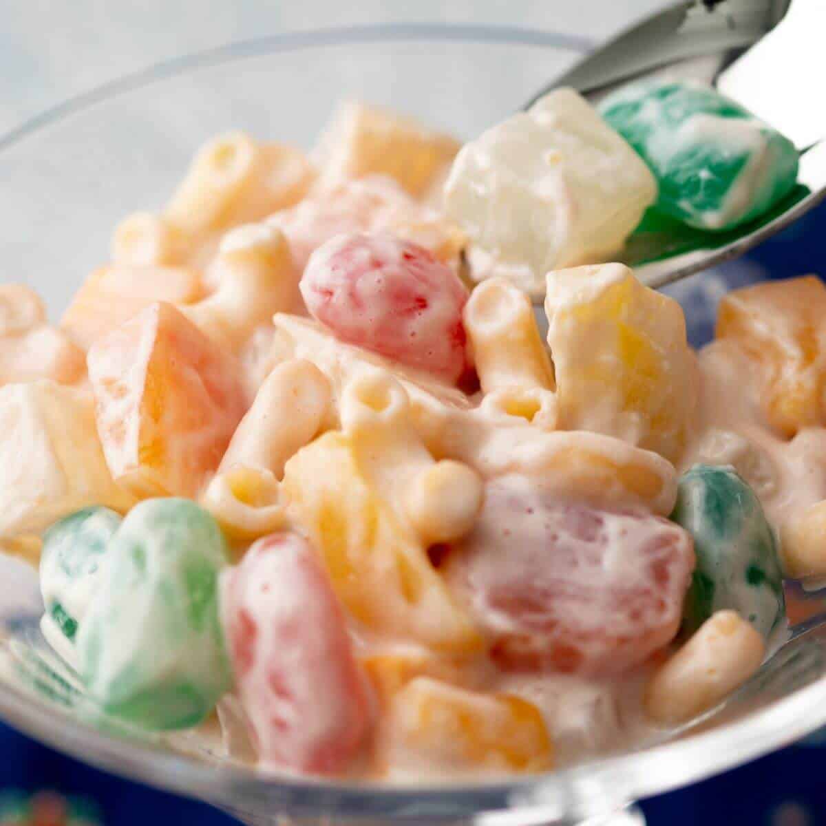 A glass bowl filled with a colorful fruit salad mixed with creamy dressing and a spoon beside it.