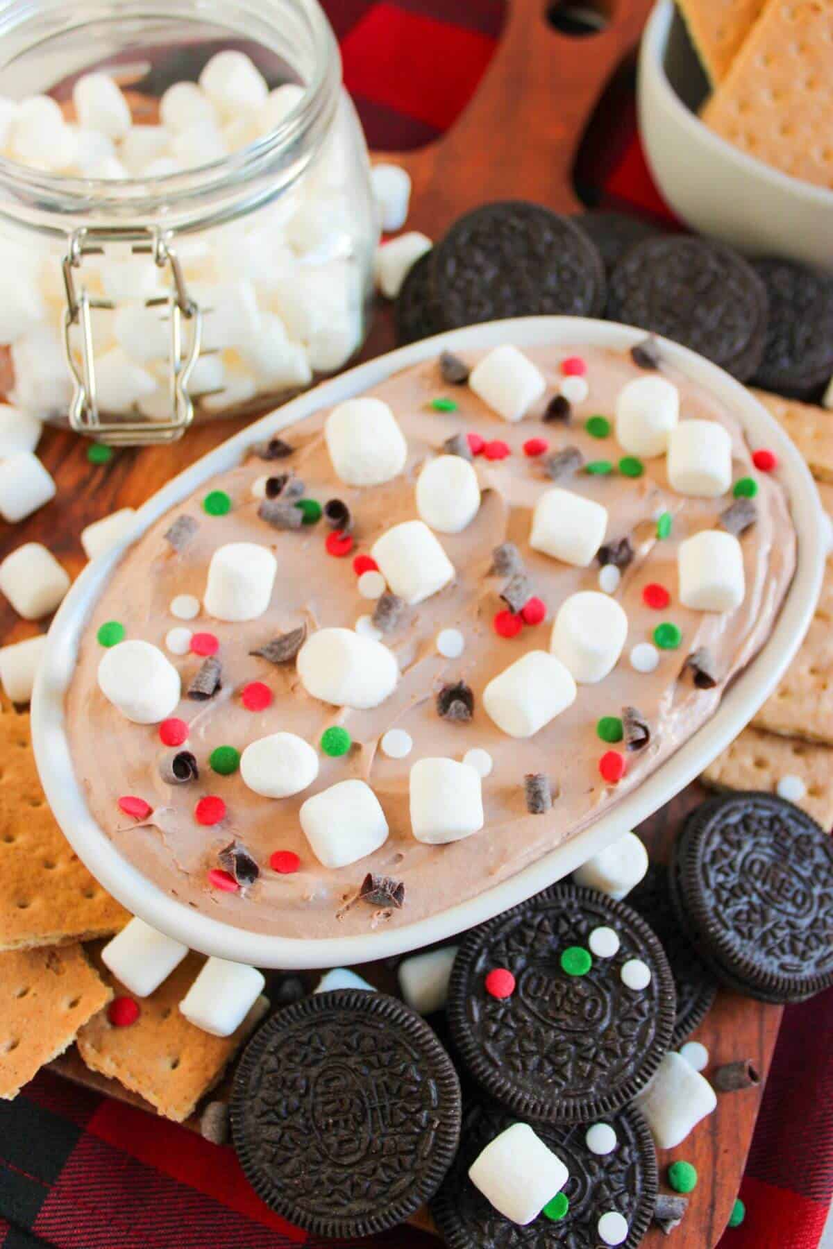 An oval dish of holiday-themed dessert dip, topped with marshmallows and sprinkles, surrounded by graham crackers, sandwich cookies, and more marshmallows in a jar.
