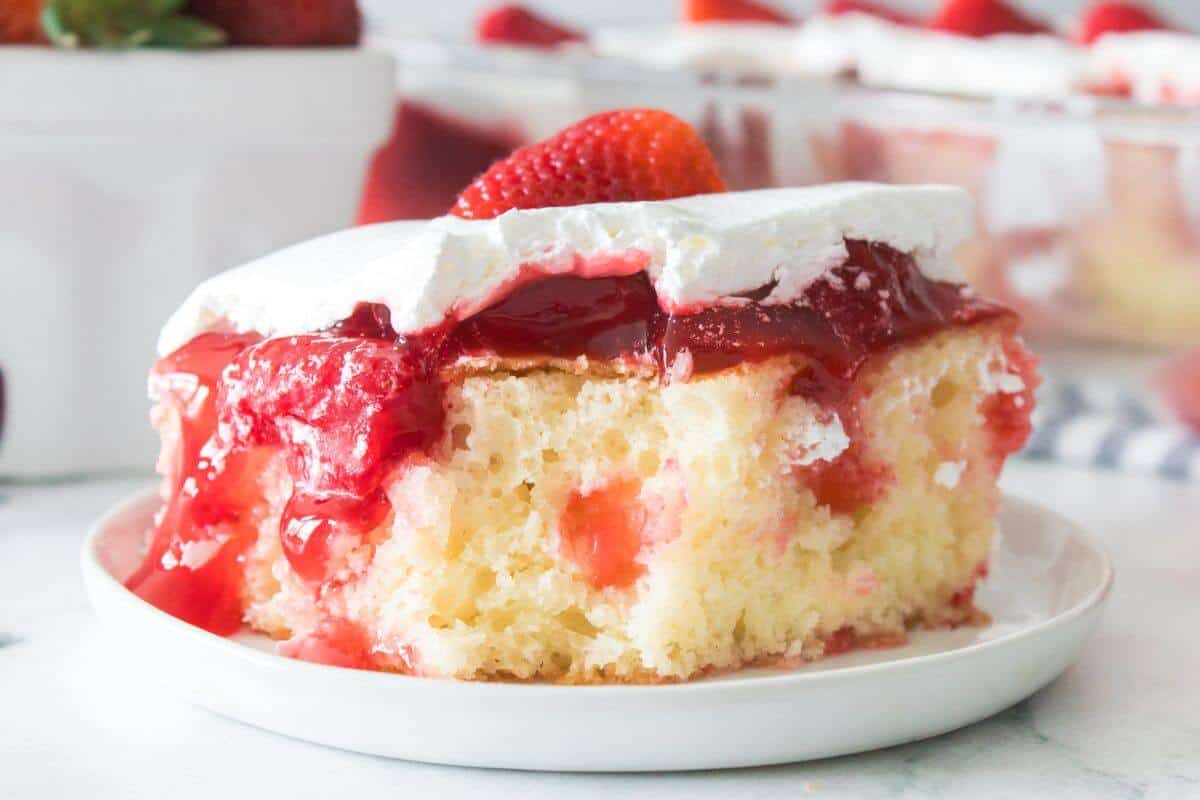A slice of strawberry cake with whipped cream topping and a strawberry garnish on a white plate.