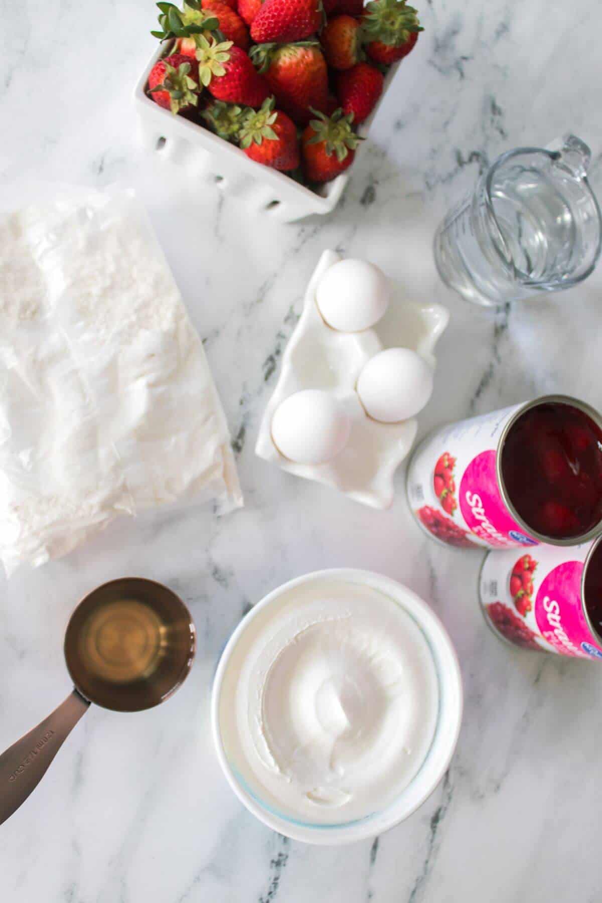 Baking ingredients on a marble countertop: strawberries, eggs, phyllo dough, measuring cup with liquid, whipped cream, and two cans of fruit filling.
