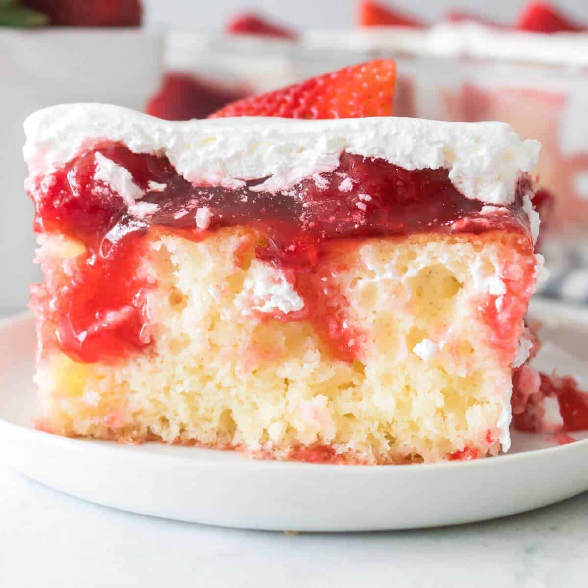 A slice of strawberry poke cake on a white plate, topped with whipped cream and strawberry sauce, with a visible moist texture.