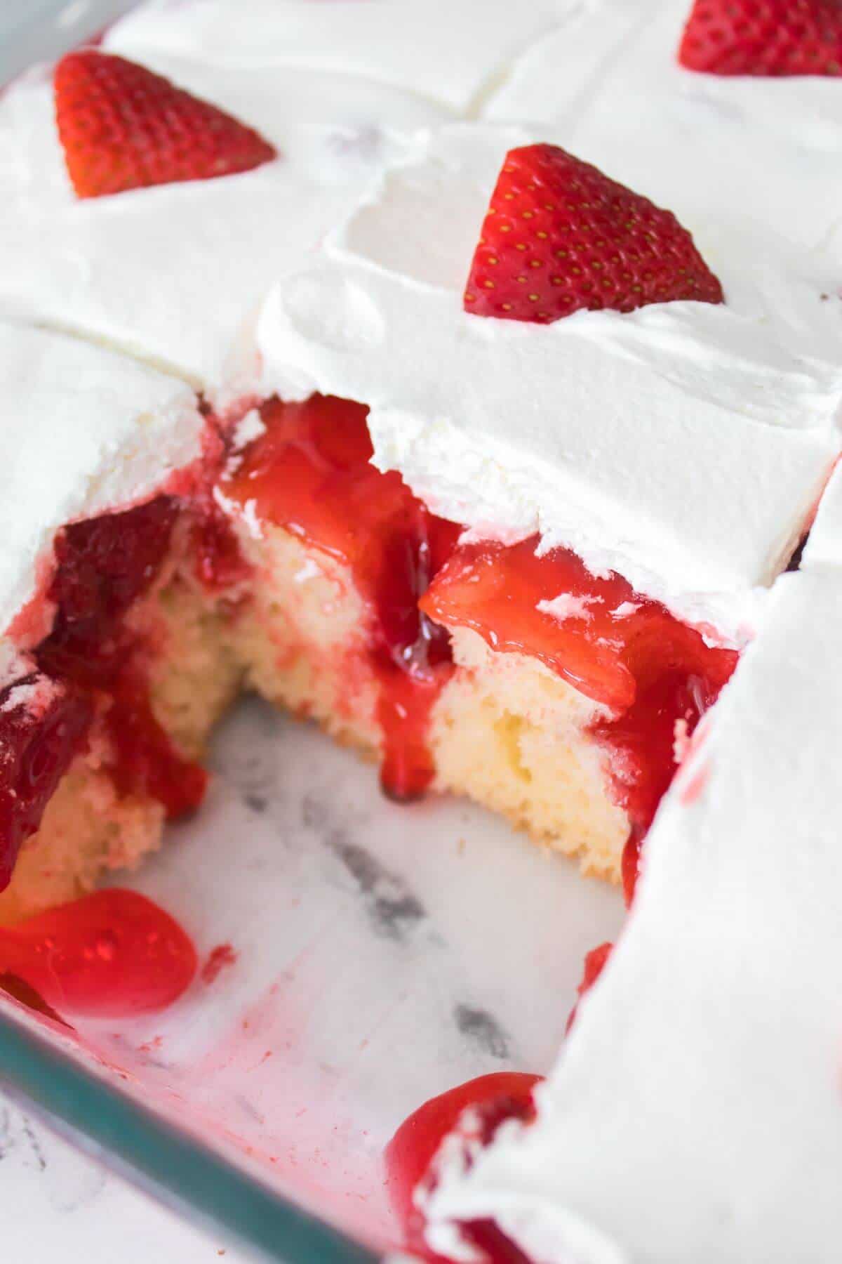 A sliced strawberry shortcake with white frosting, topped with strawberry halves, in a glass dish on a marble surface.