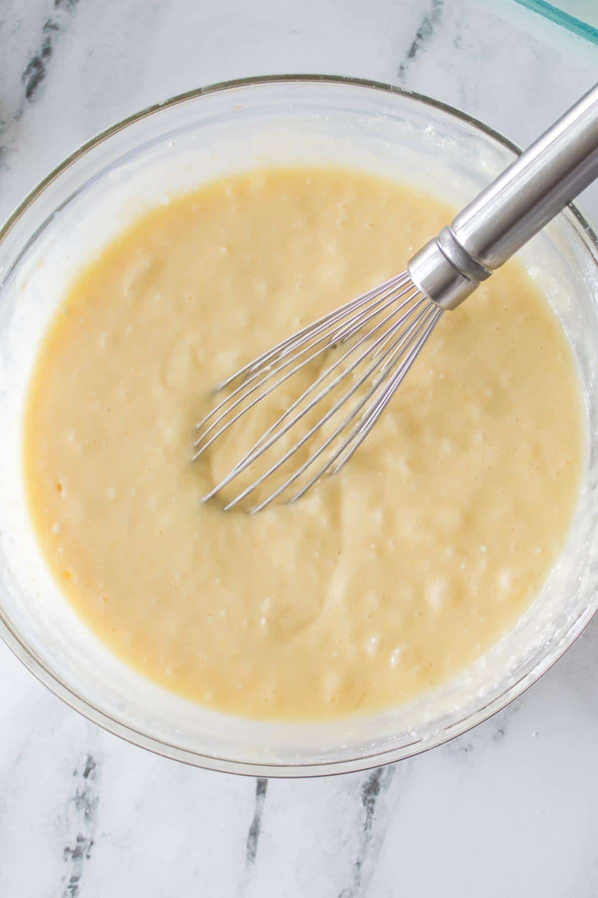Bowl of banana bread batter with a metal whisk on a white marble countertop.