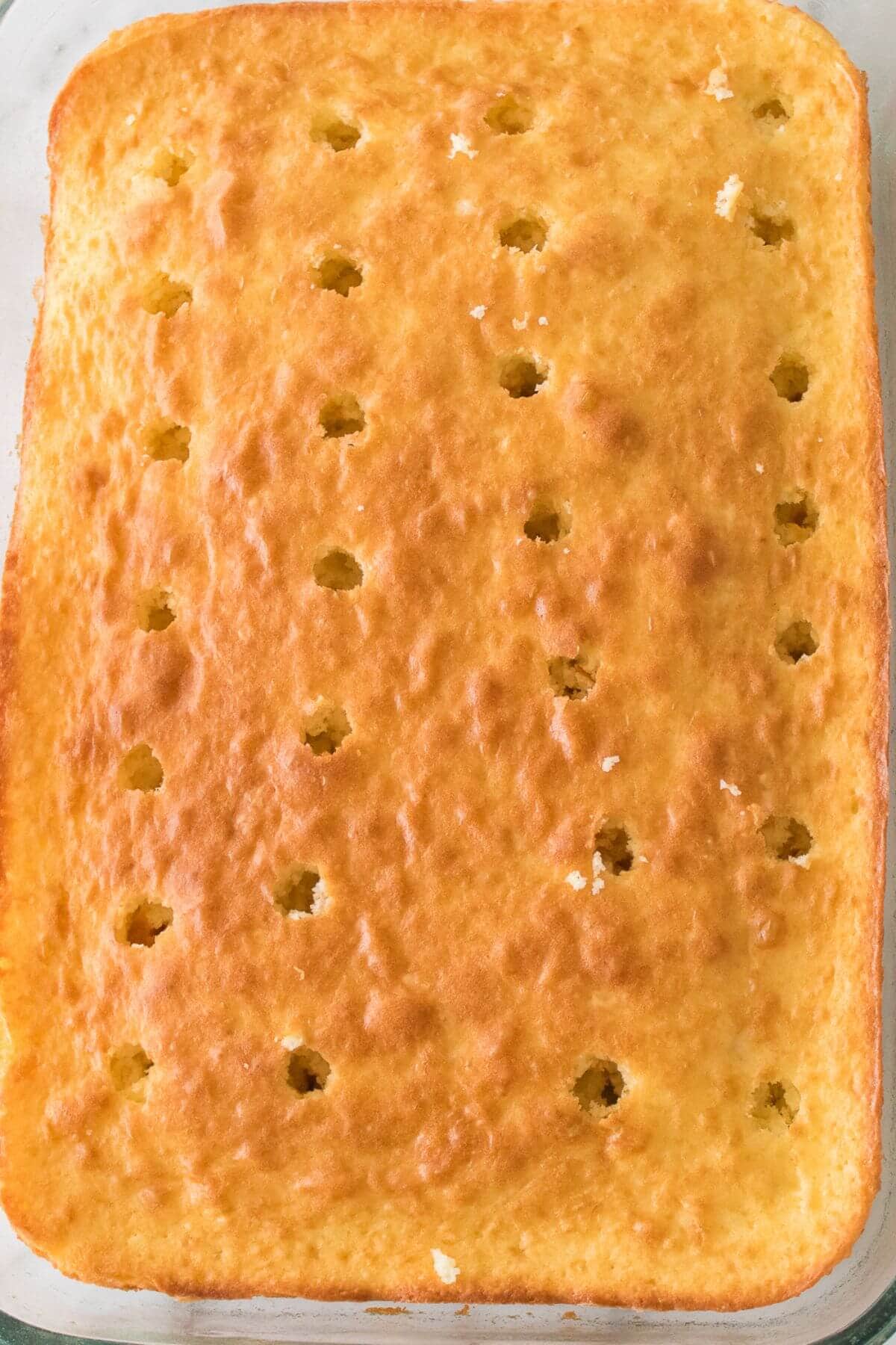 A rectangular cake with a golden-brown top has evenly spaced holes poked into its surface.