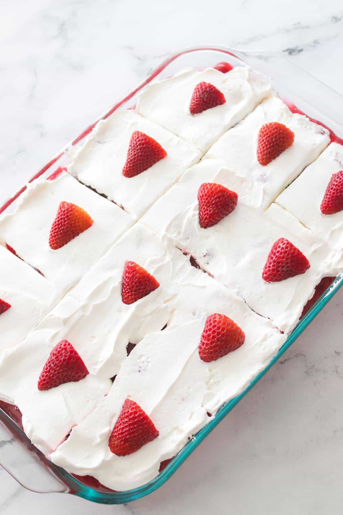 A glass dish with a layered dessert topped with whipped cream and whole strawberries, divided into rectangular portions.
