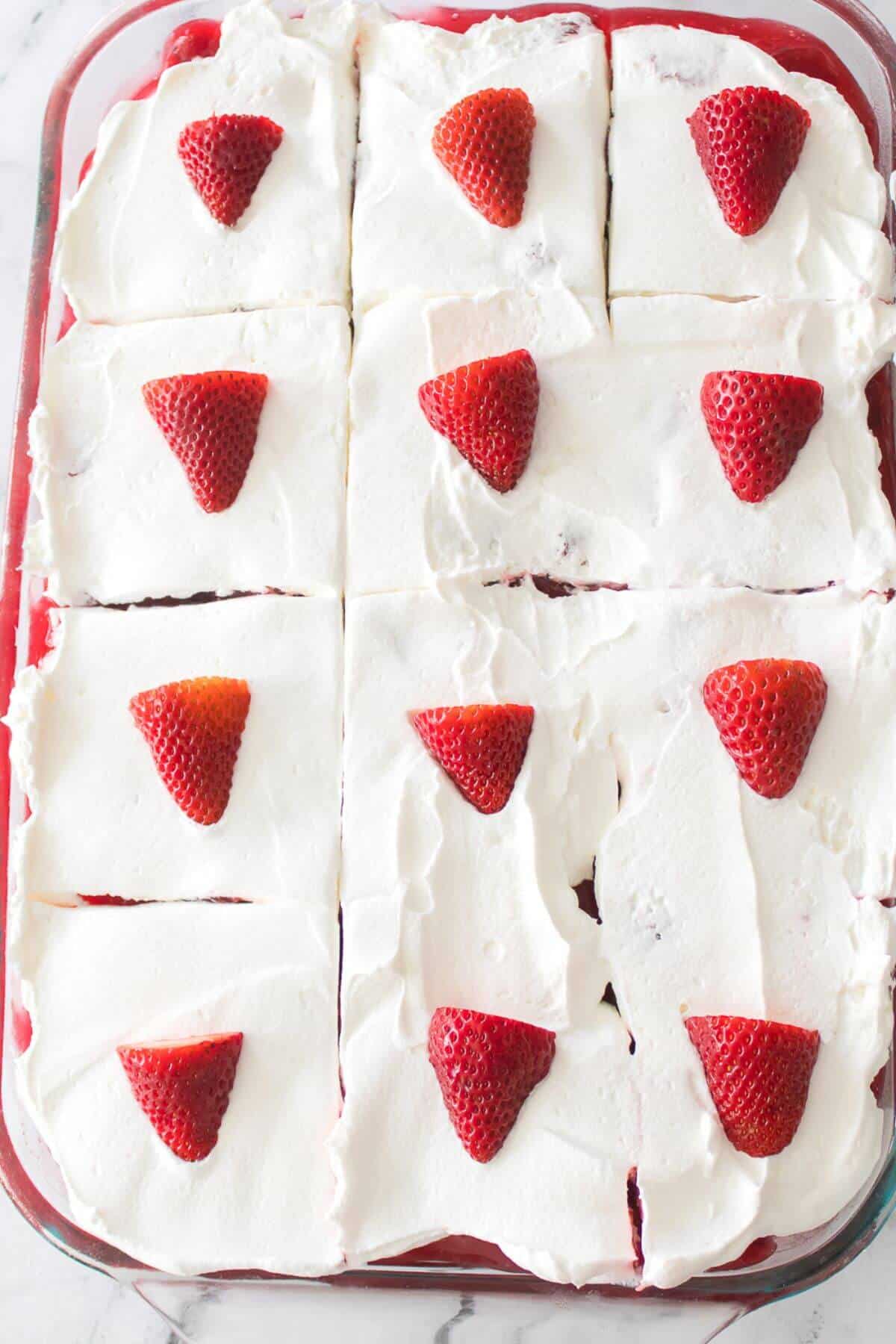 A dessert with nine squares of cream-topped layers, each adorned with a sliced strawberry, in a glass dish.